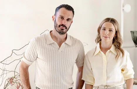 A man and woman standing indoors in front of a white wall with a decorative plant.