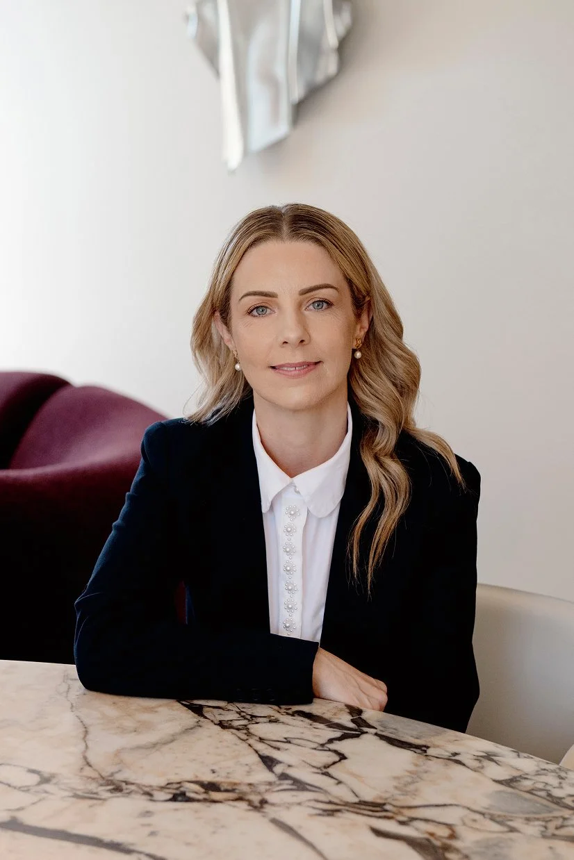 A woman with light brown, wavy hair and blue eyes sitting at a marble table, wearing a black blazer and a white shirt with pearl earrings, in a room with white walls and a maroon chair in the background.