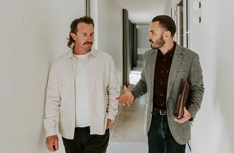 Two men walking and talking in a hallway, one holding a folder.