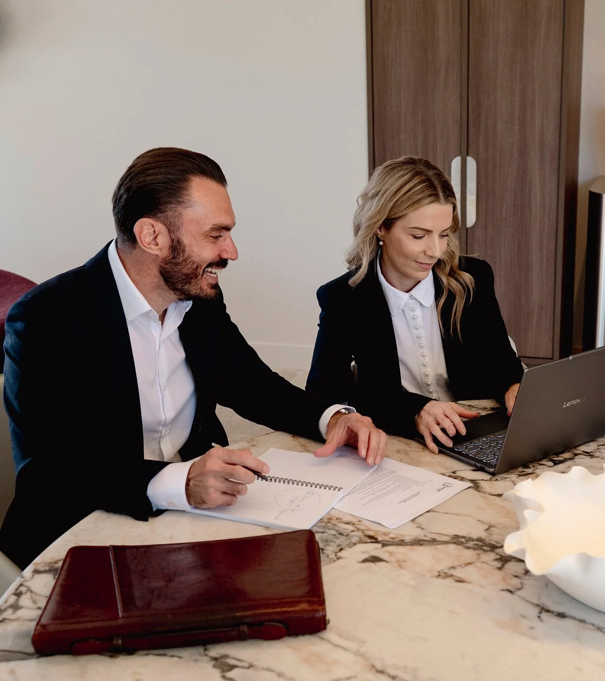 Two business professionals, a man and a woman, sitting at a marble table, smiling and working together with documents and a laptop.