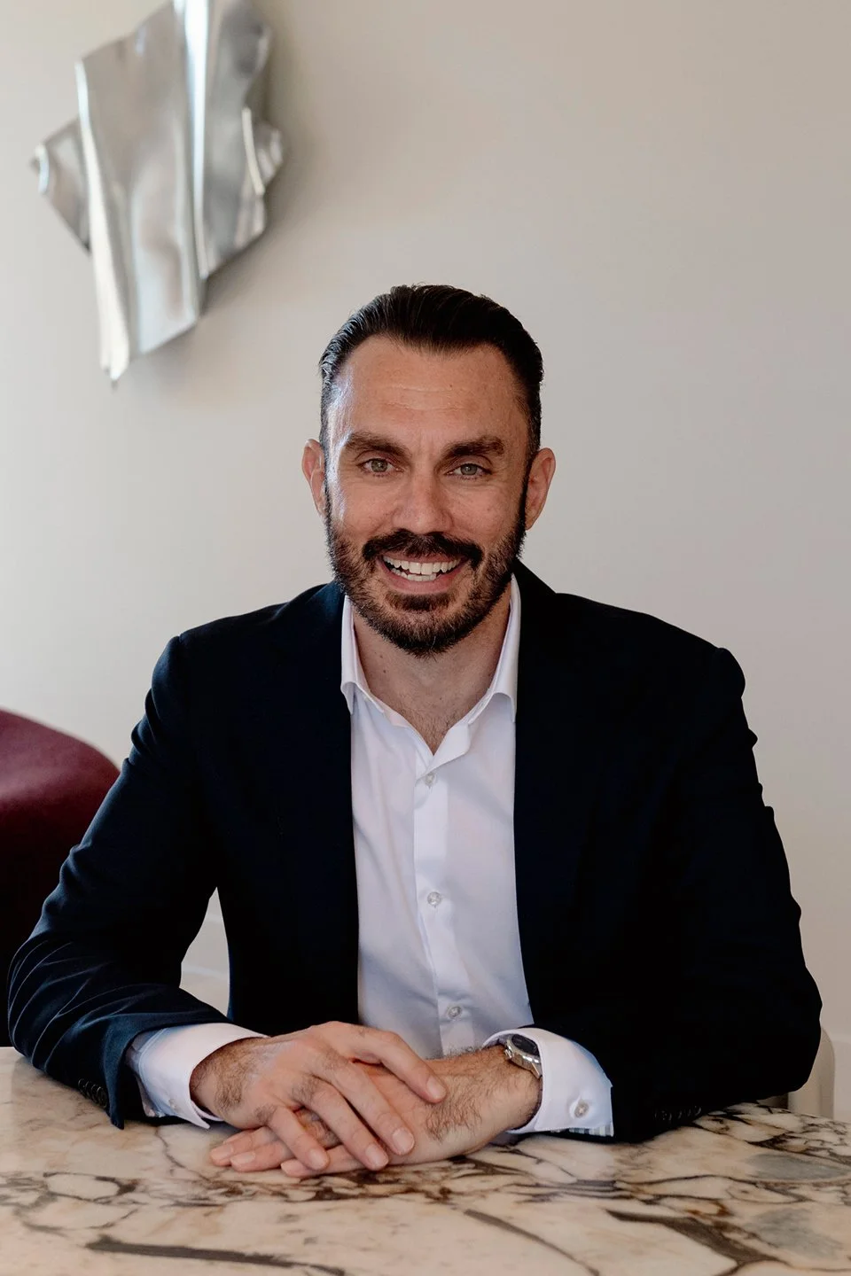 A man with dark hair and a beard, wearing a black suit and white shirt, smiling and sitting at a marble table.