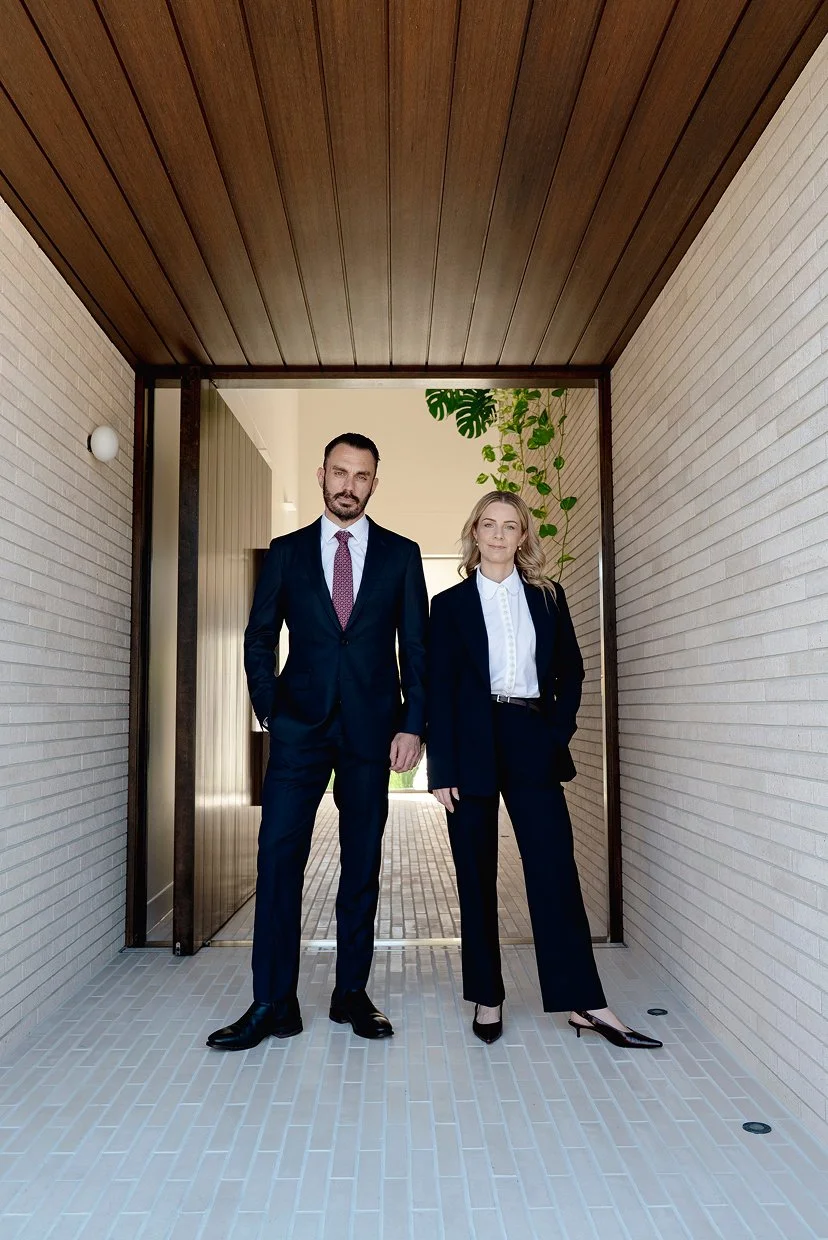 Two professionally dressed people, a man and a woman, standing in a modern, covered outdoor corridor with white brick walls and a wooden ceiling.