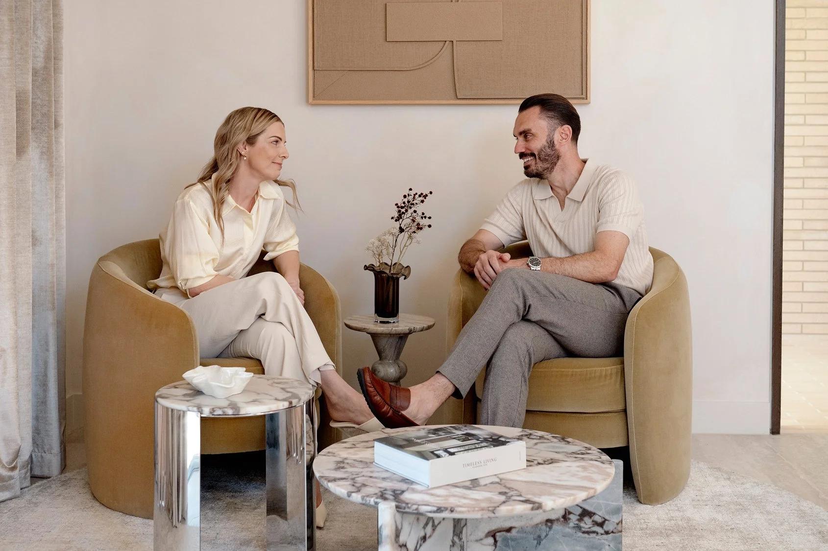 A man and woman sitting in beige armchairs facing each other in a modern, minimalistic room. They are engaged in a conversation with a small table in between, holding a decorative vase and a stack of books. The woman is wearing a cream-colored outfit, and the man is dressed in a light shirt and gray pants.
