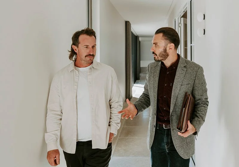 Two men having a serious conversation in a hallway, one in casual attire and the other in a blazer holding a folder.