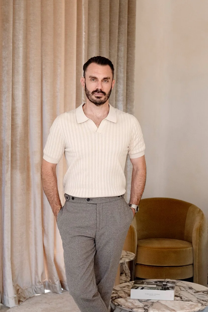 A man with dark hair and beard, wearing a cream collared shirt and gray pants, standing in a room with beige curtains and a brown armchair.