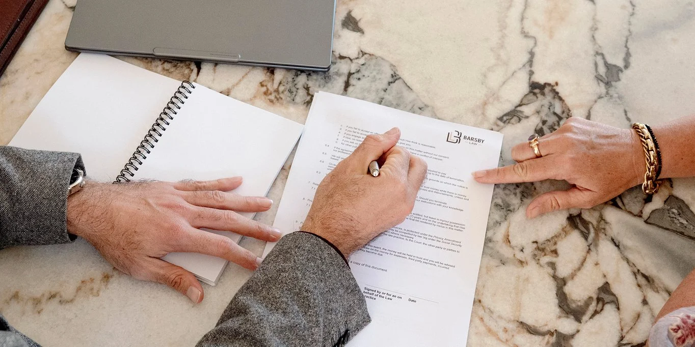 Two people reviewing and signing legal documents on a marble table, with a closed laptop and notebooks nearby.