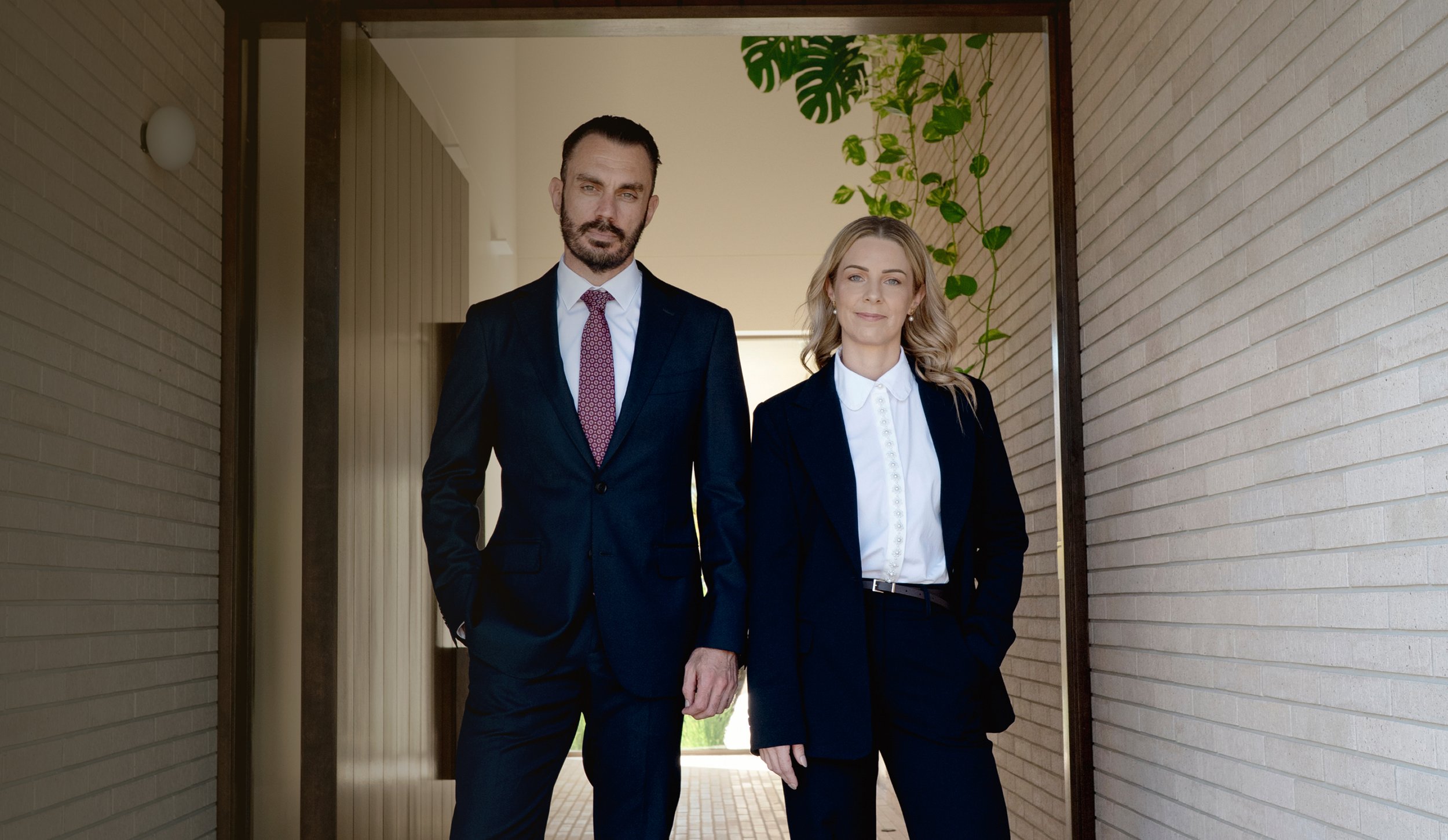 A man and woman in formal business attire walking together outdoors near a brick building, holding hands.