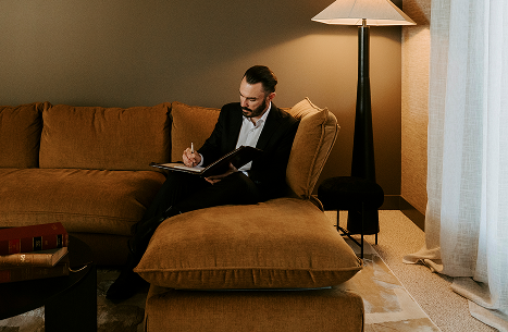 Man in black suit sitting on a large brown sofa, writing in a notebook with a coffee table and a tall lamp nearby in a living room.