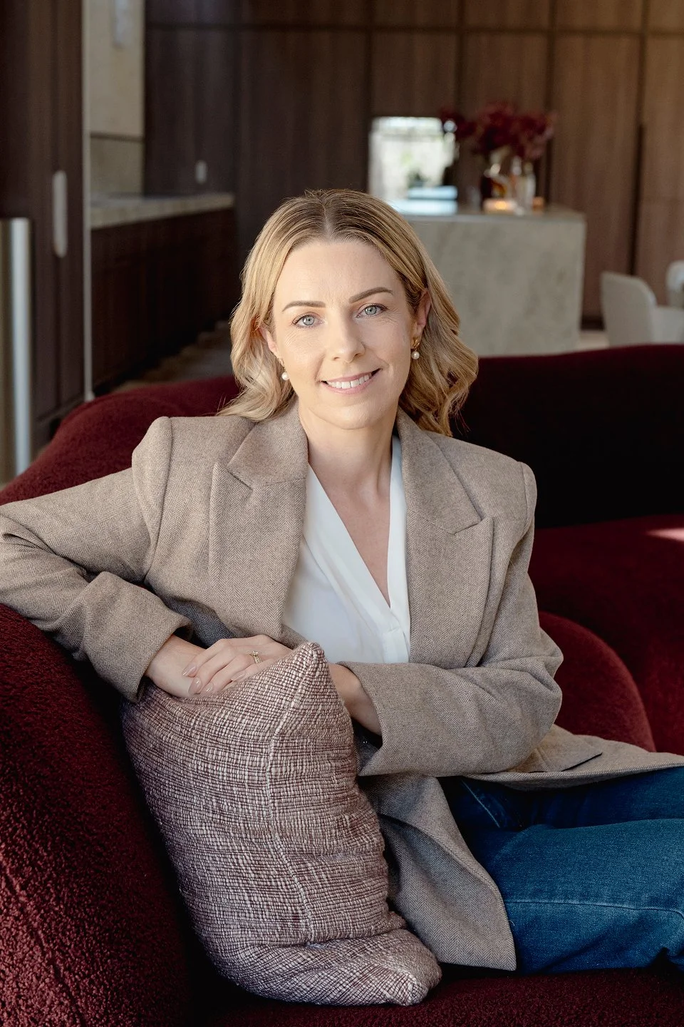 A woman with shoulder-length blonde hair, wearing a beige blazer over a white top, is sitting on a red sofa with a patterned throw pillow in a modern living room.