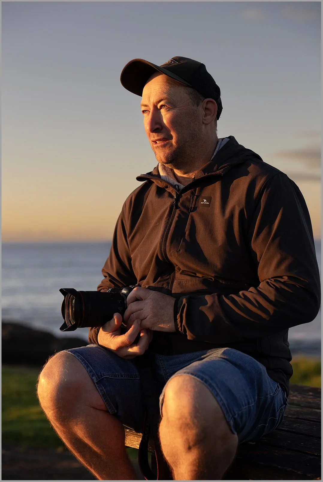 A man in a black jacket, shorts, and a cap sitting outdoors at sunset, holding a camera and looking into the distance.