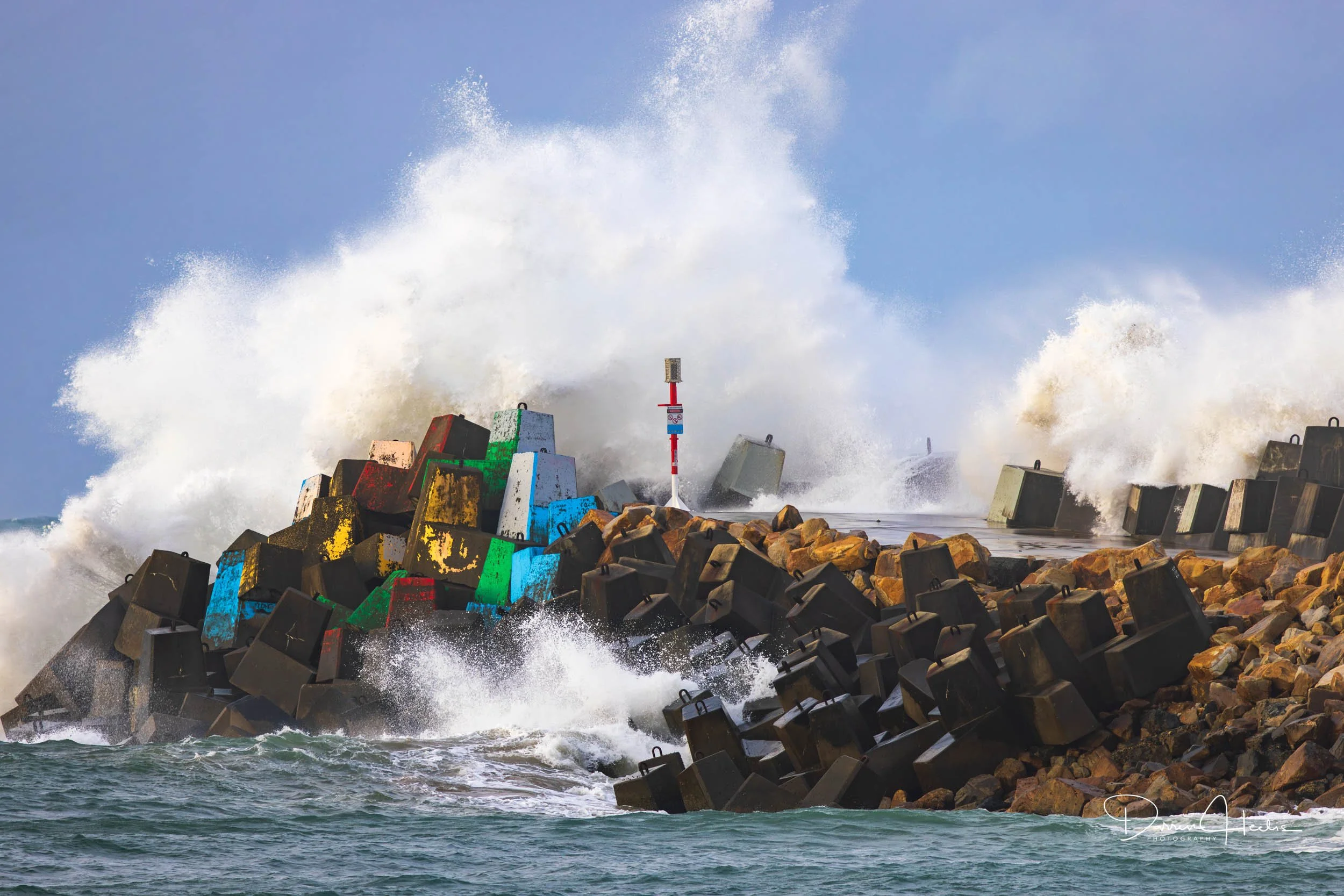 Waves crashing against a rocky breakwater with large concrete blocks, with water splashing high into the air against a clear blue sky.