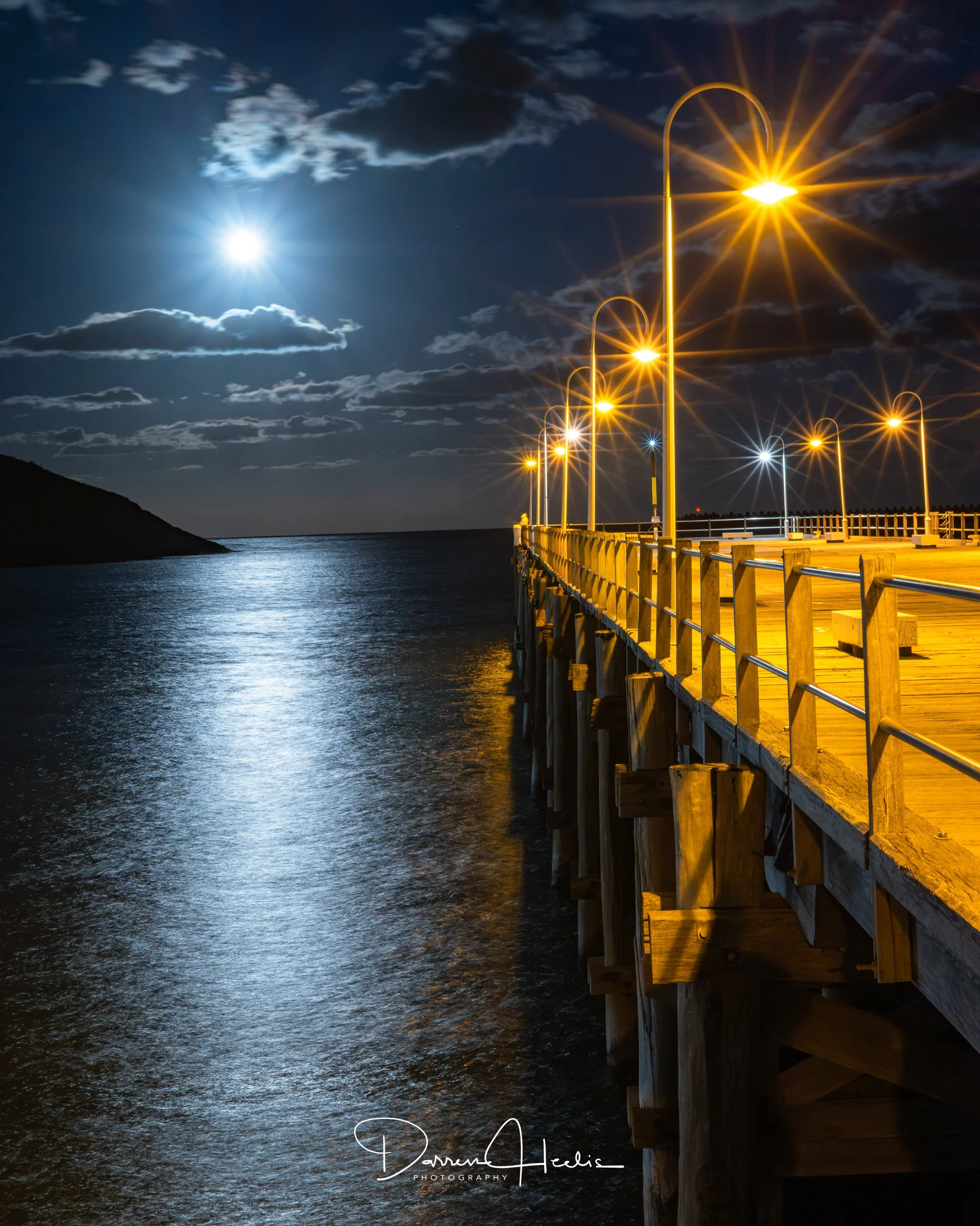 Night scene of a moonlit sky over a pier with wooden railings and street lamps casting warm light, reflected on the water below, with clouds illuminated by the moon.