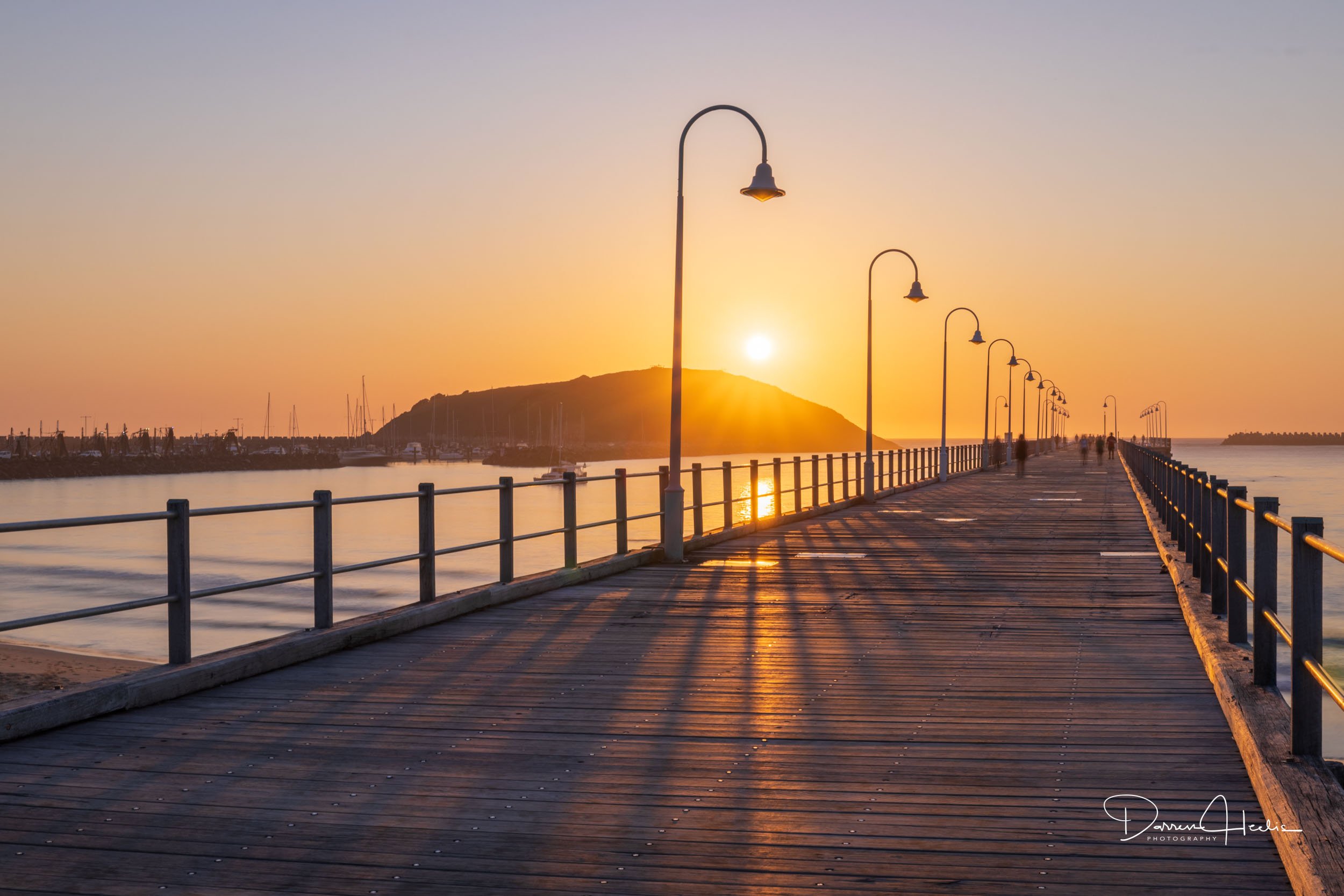 A wooden pier with lampposts extending into the ocean at sunset, with a hill and boats in the background.