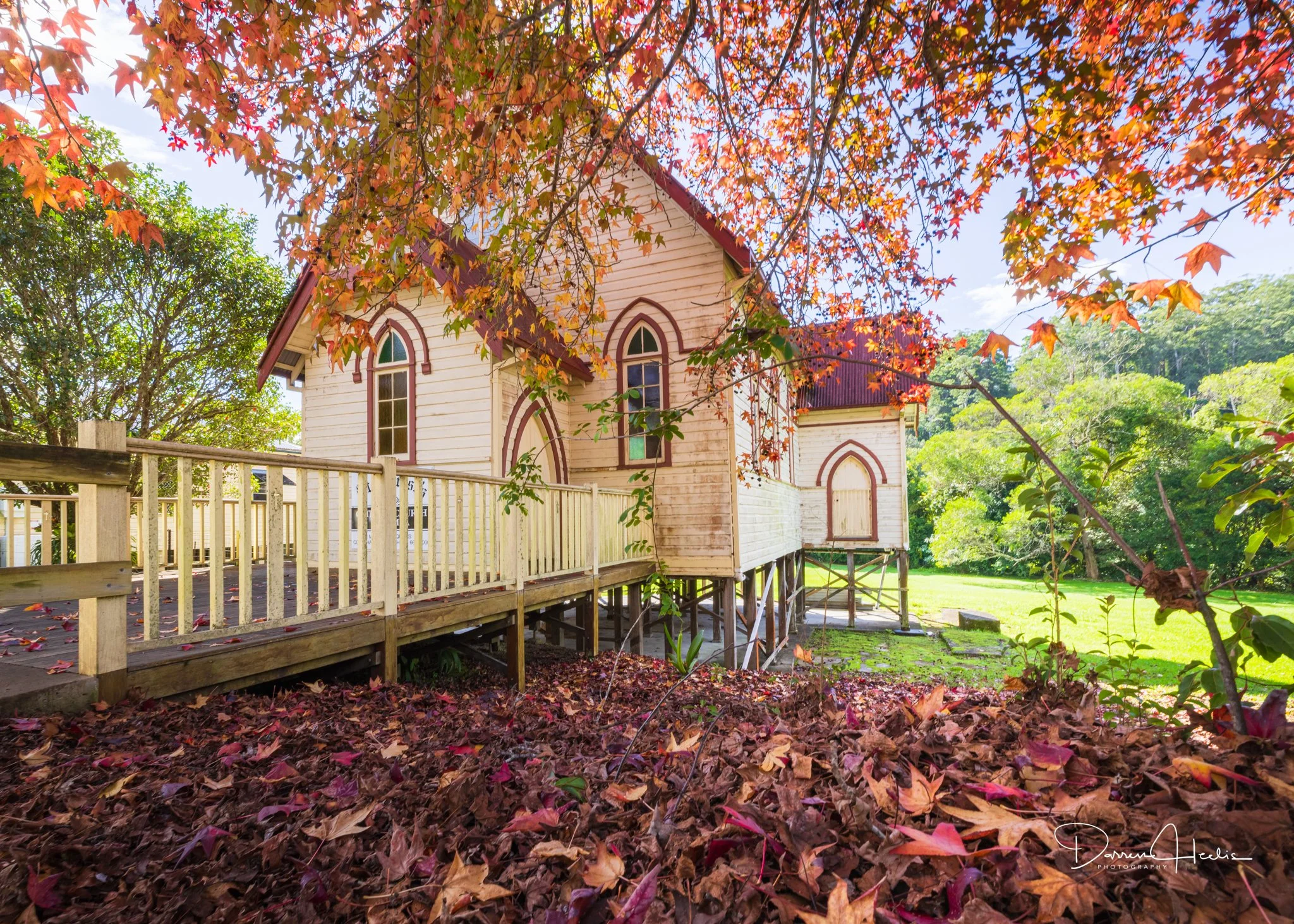 A white wooden church with Gothic-style arched windows, surrounded by trees with colorful autumn leaves and a wooden fence in the foreground.