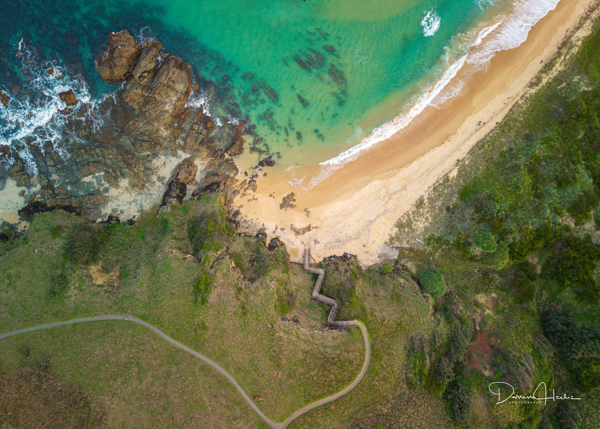 An aerial view of a beach with rocks, turquoise water, sandy shore, and greenery on the cliffs, with a trail winding along the landscape.