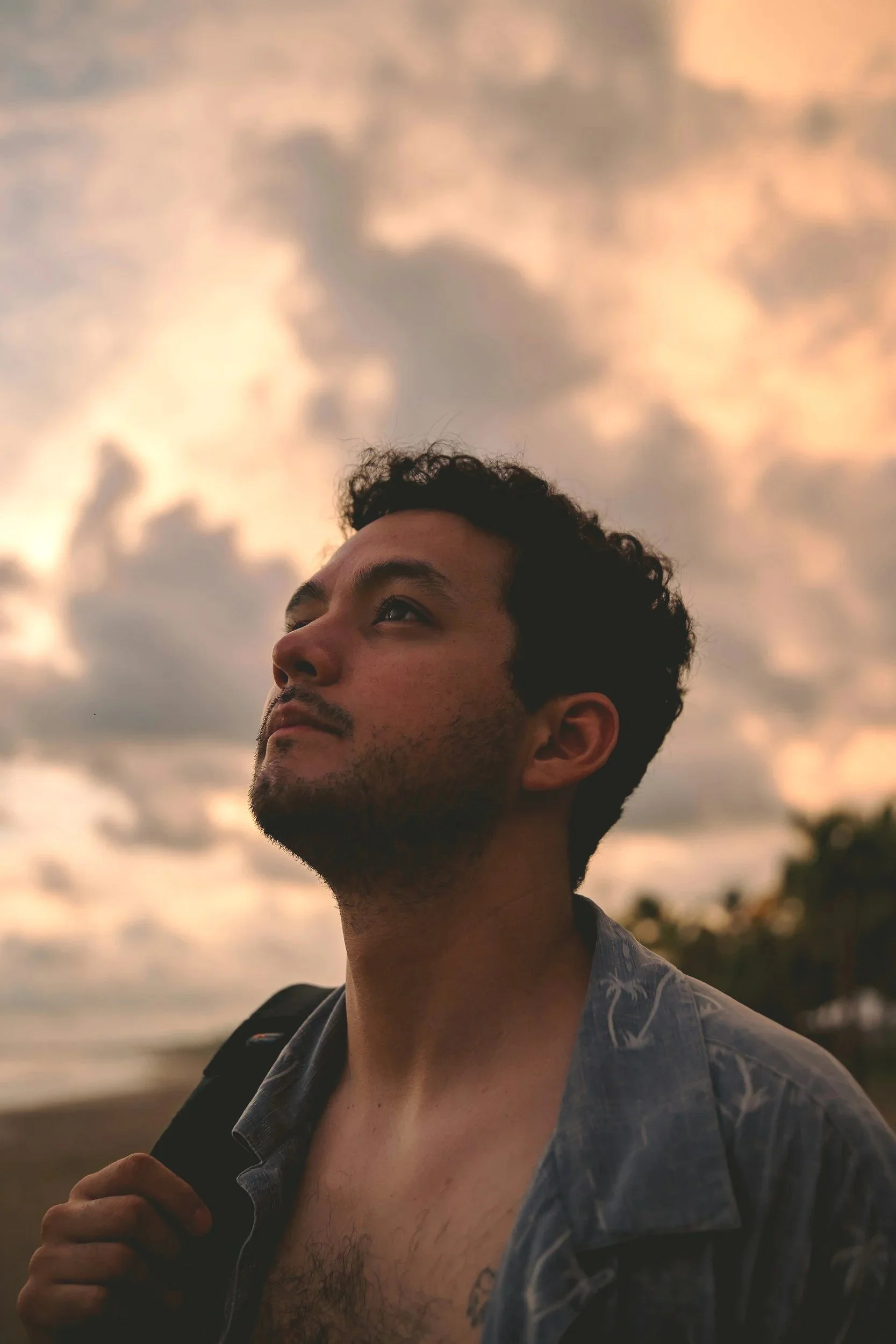 A man with short, curly black hair and light facial hair looking up against a cloudy sky at sunset, wearing a denim shirt and carrying a backpack.