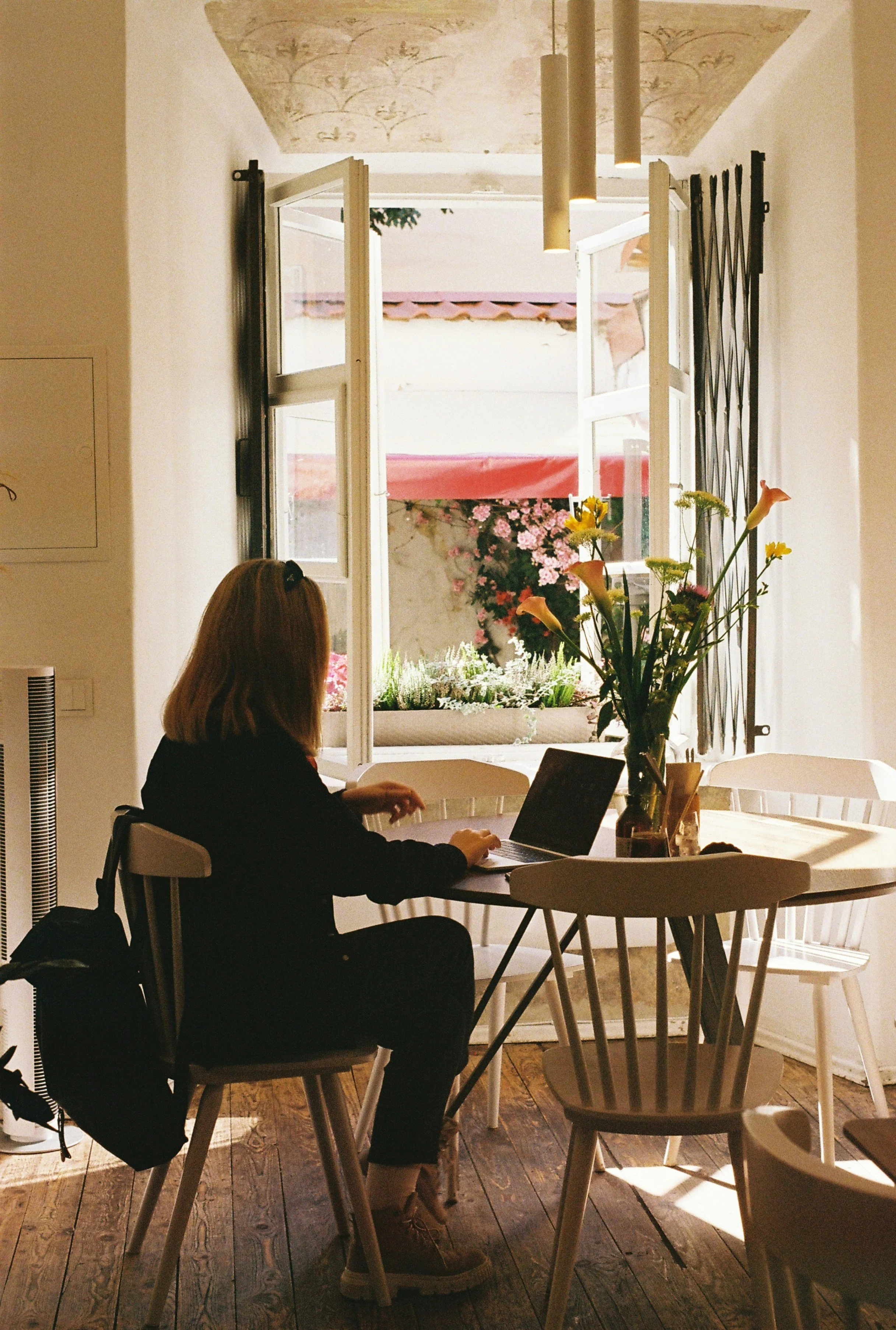 A woman with red hair wearing a black sweater and beige boots, sitting at a round white table with a laptop and a backpack, in a bright room with open windows, flowers on the table, and sunlight streaming in.