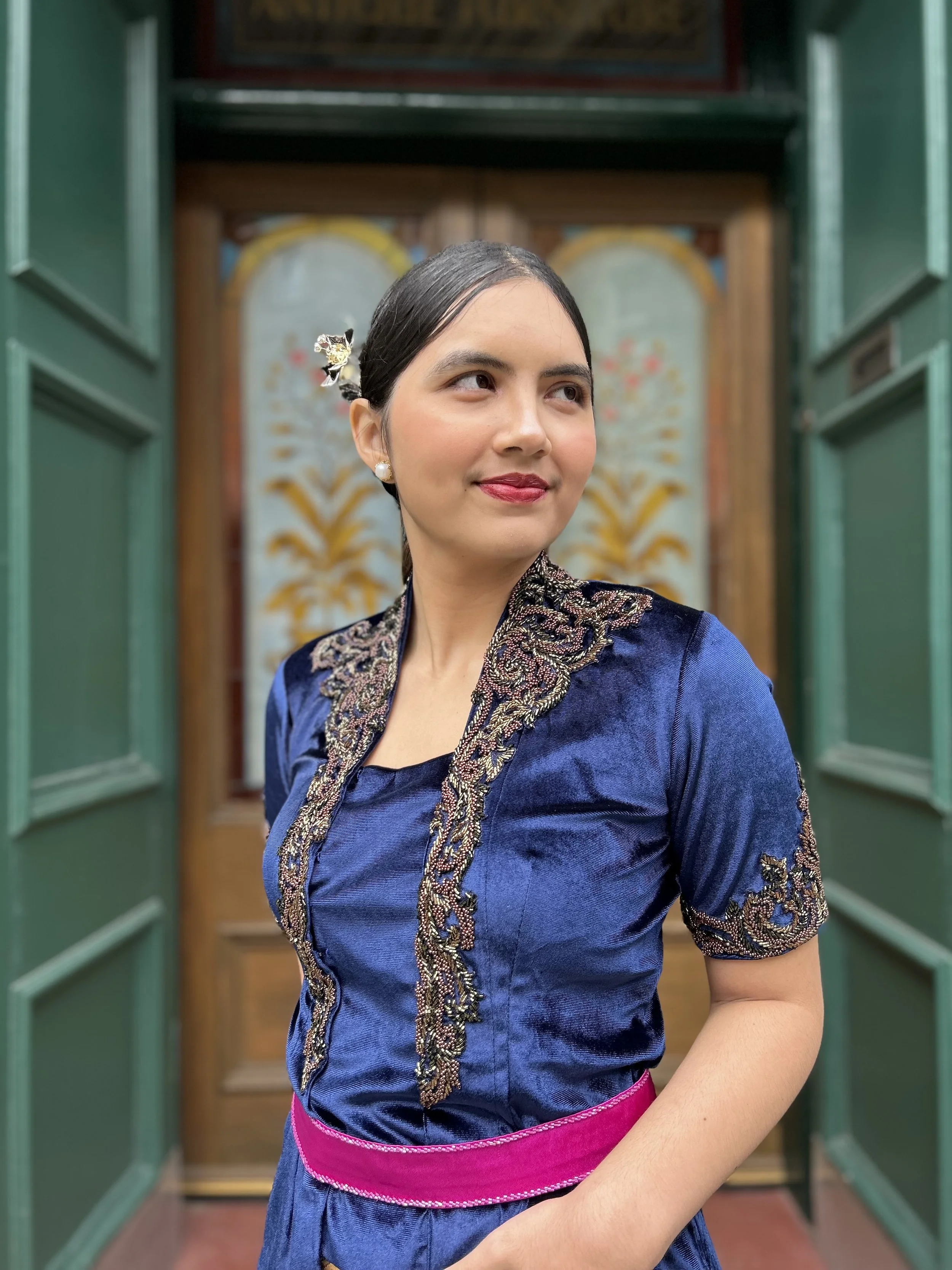 A woman wearing a traditional dark blue dress with intricate gold embroidery, standing at an open green door with a wooden door and decorative wall behind her, and a pink sash around her waist.