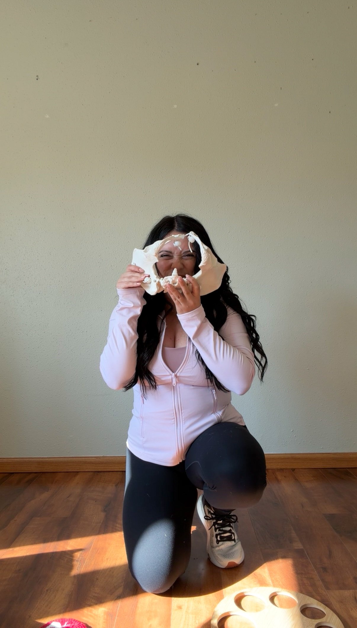 Woman kneeling on one knee, wearing black leggings, white sneakers, and a pink zip-up jacket, holding a white foam ring around her face, laughing, indoors with a plain yellow wall and hardwood floor.
