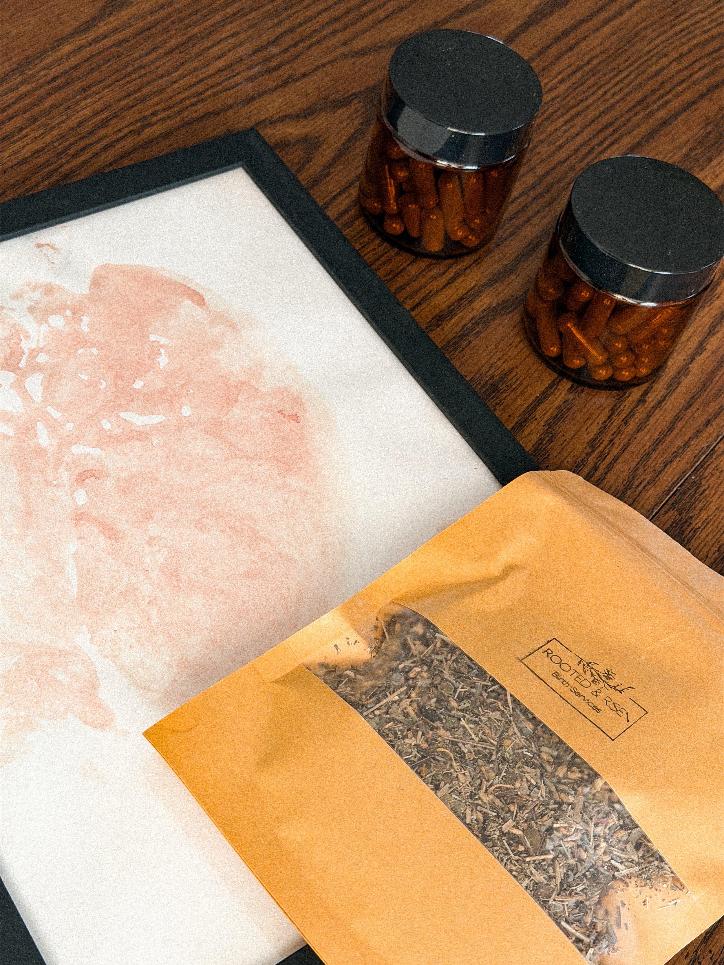 Two jars of capsules on a wooden table, a partially visible artist's painting, and a package labeled "Rooted & Rise: Earth, Sun, Water" containing dried herbs or plants.