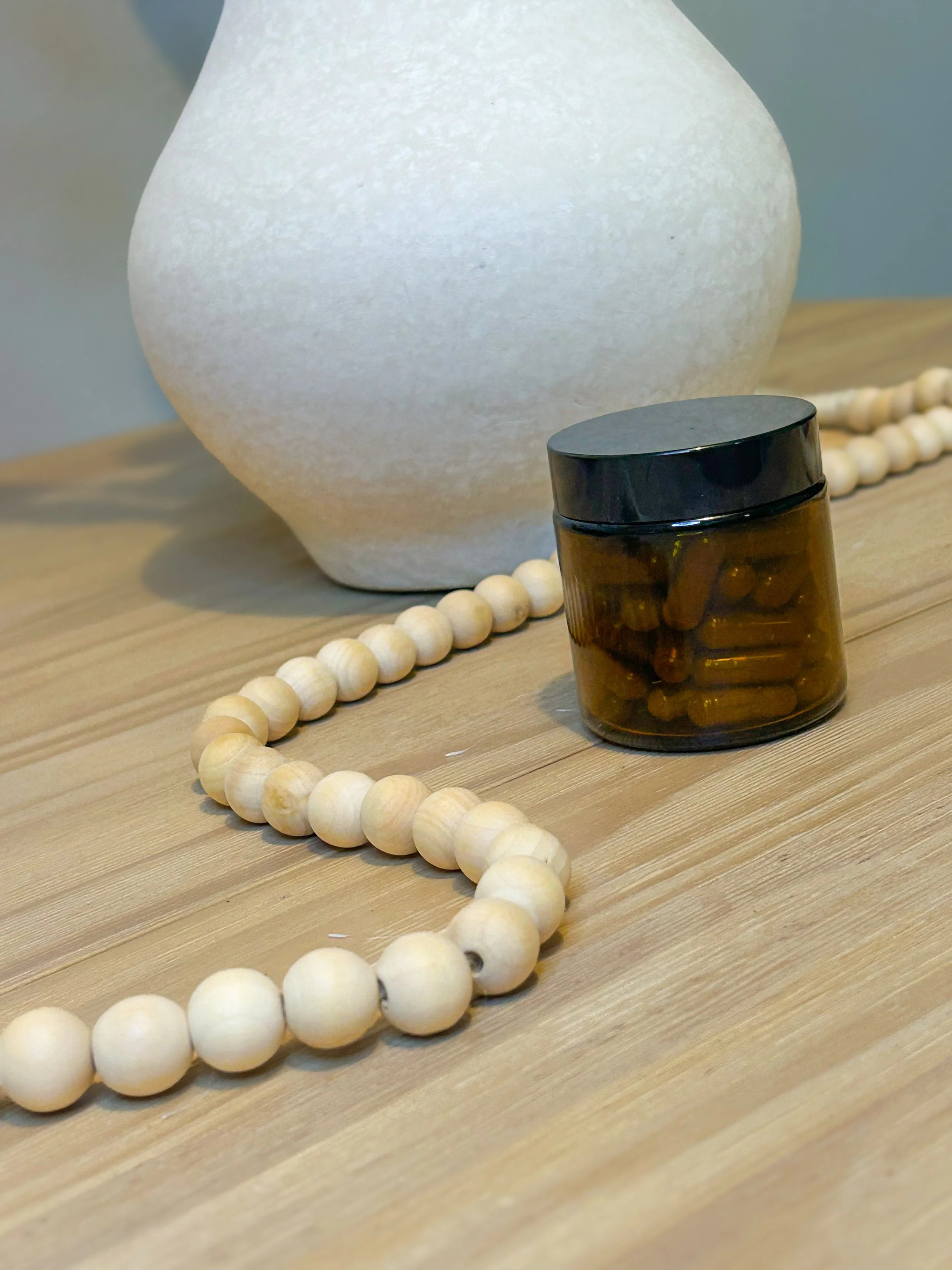 A white decorative vase, a brown glass container with capsules, a beaded necklace, and a wooden surface.