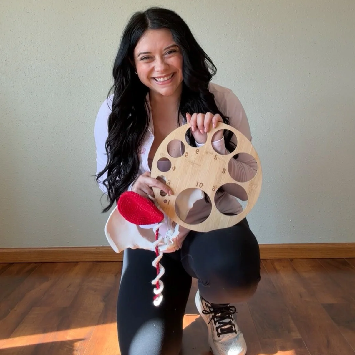 A woman with long black hair smiling and kneeling on the floor, holding a wooden shape sorter toy with holes labeled with numbers, and a red and white plush toy draped over her arm. She is wearing a beige jacket, black pants, and white sneakers, with a plain wall in the background.