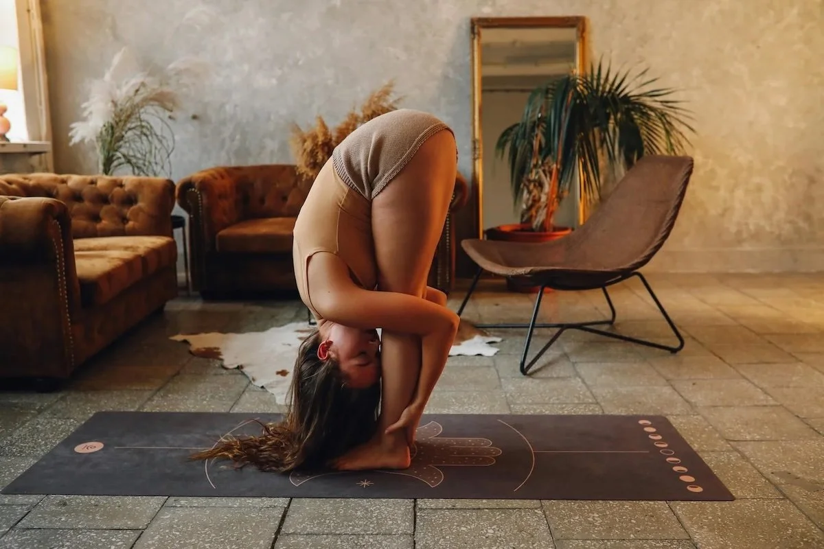 A woman practicing yoga in a rustic living room, performing a forward bend pose on a black yoga mat.