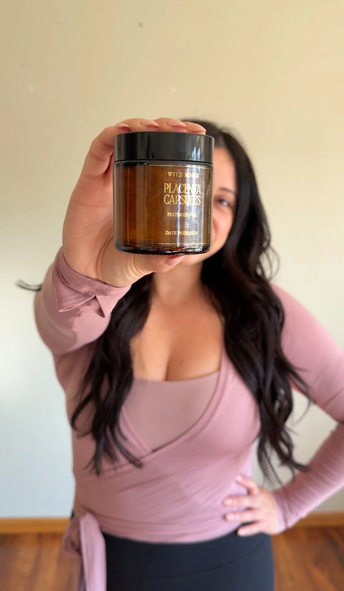 A woman with long dark hair, wearing a pink wrap top, is holding a jar labeled 'Placenta Capsules' towards the camera, partially obscuring her face.