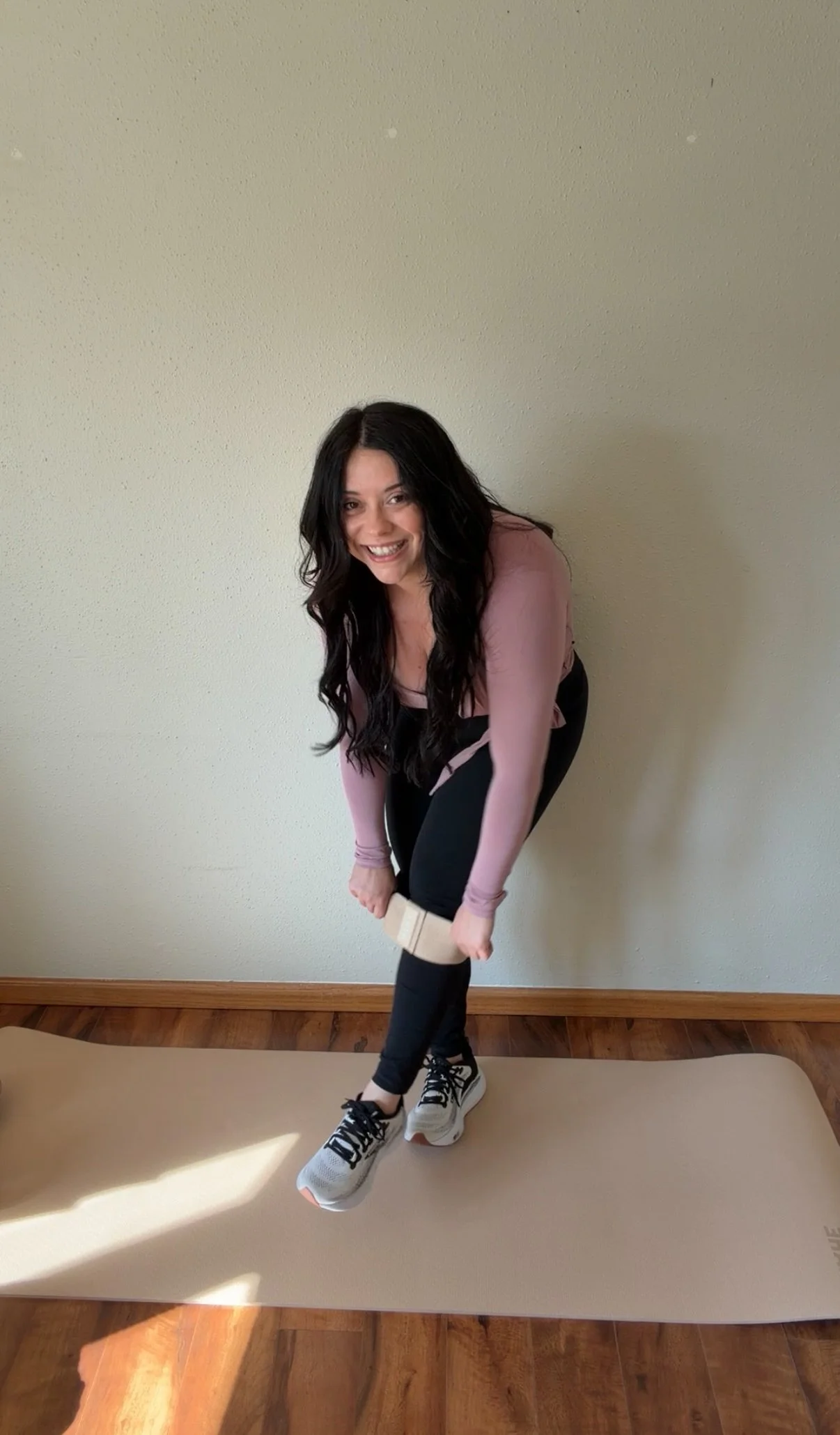 Woman in black jeans, pink long-sleeve top, white sneakers standing on an exercise mat, holding a water bottle in front of a plain wall.