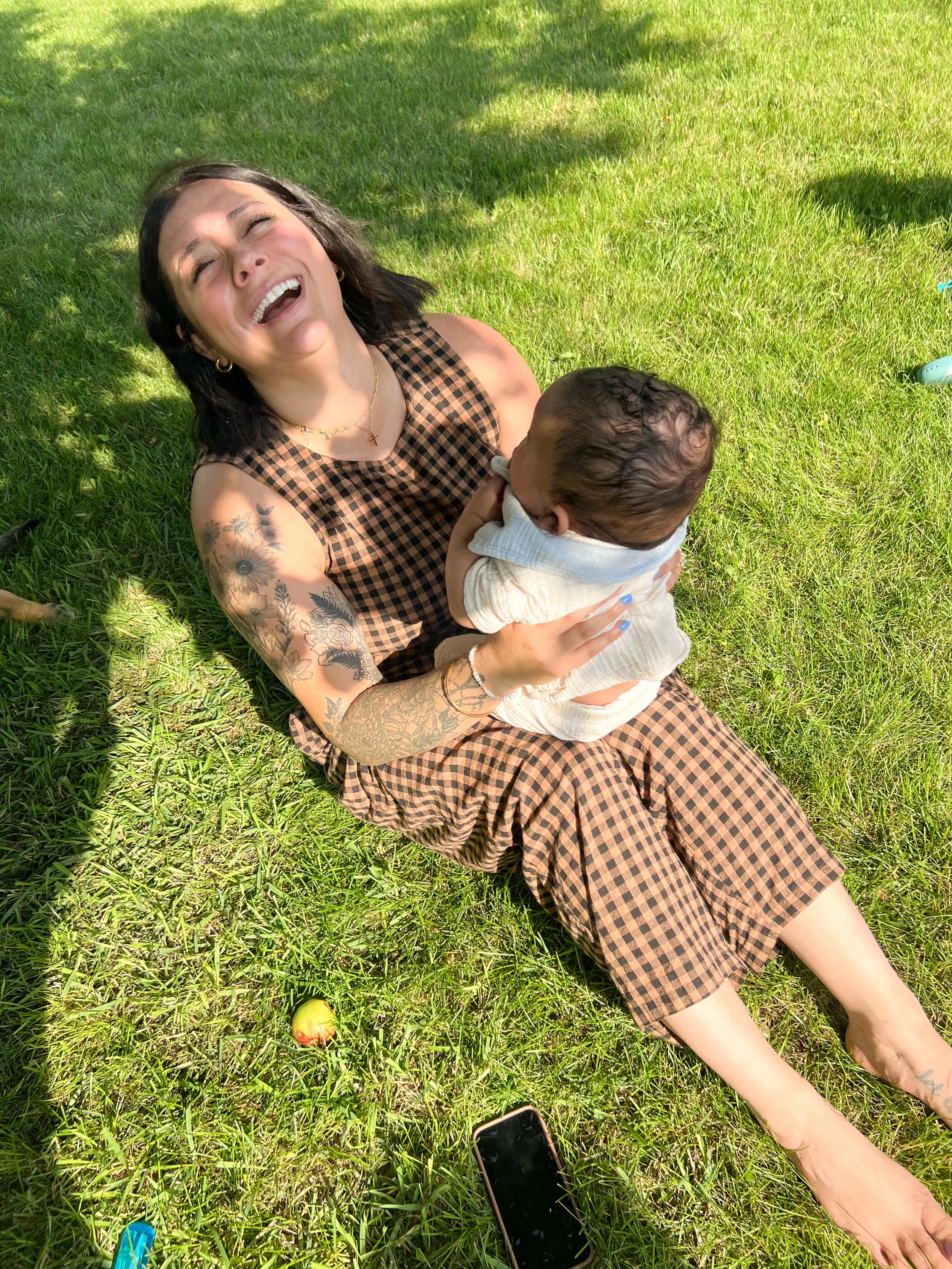 A woman with dark hair and tattoos on her arm, wearing a checkered dress, is sitting on the grass, laughing joyfully while holding a baby. They are outdoors on a sunny day with shadows cast on the grass. A smartphone is on the grass near her feet, and an apple is also visible.