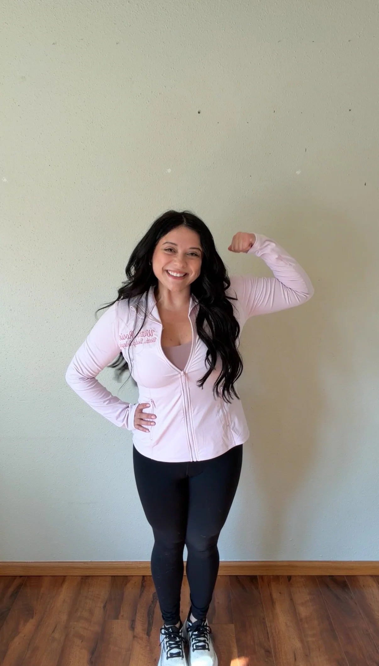 A woman with long dark hair smiling and flexing her right arm, wearing a light pink zip-up jacket, black leggings, and athletic shoes, standing inside on a wooden floor against a plain light-colored wall.
