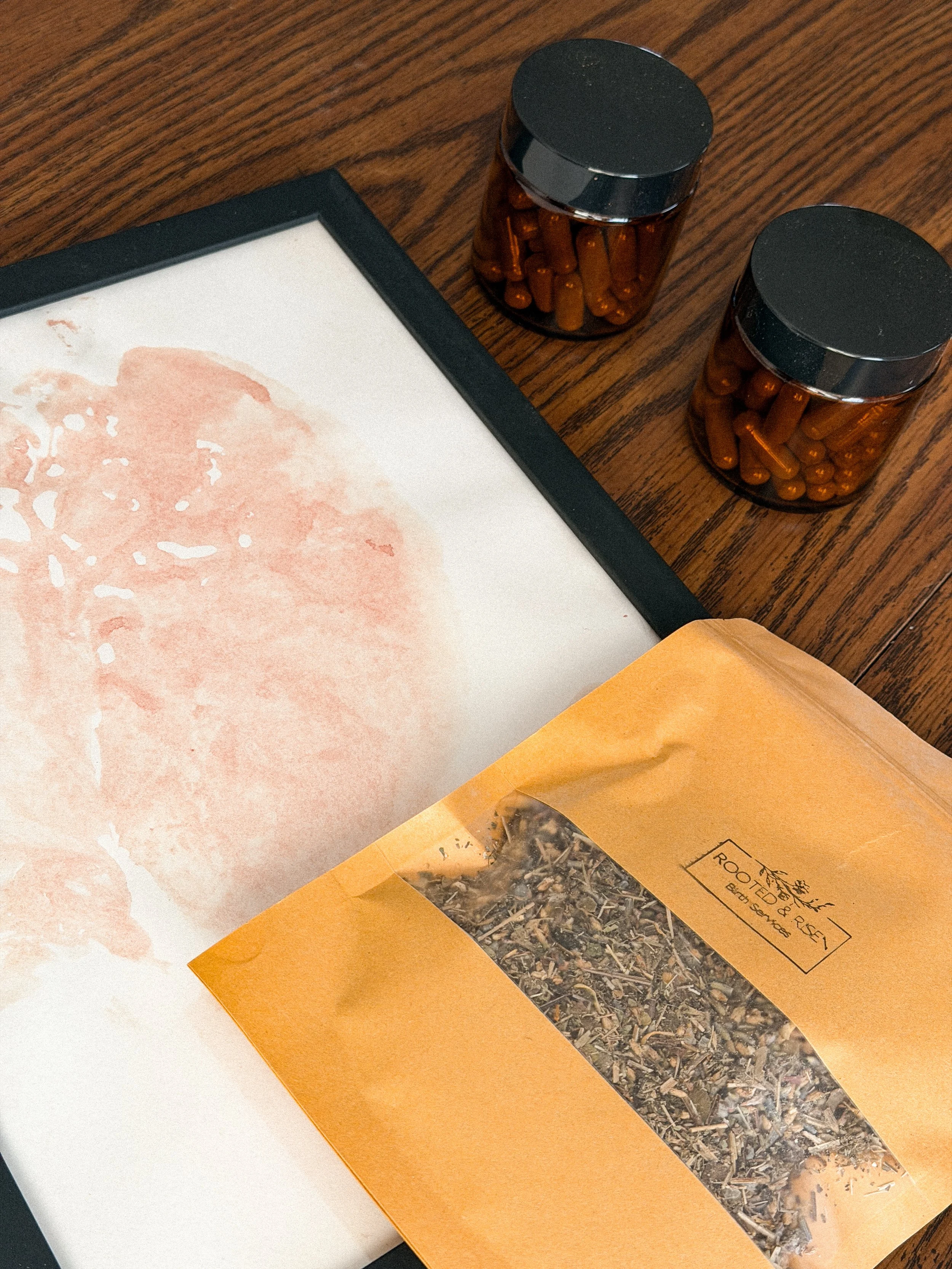 Two small amber-colored pill bottles with black caps on a wooden surface, next to a package of dried herbs labeled 'ROOTED & RISE! Earth Surfaces' on an orange paper bag.