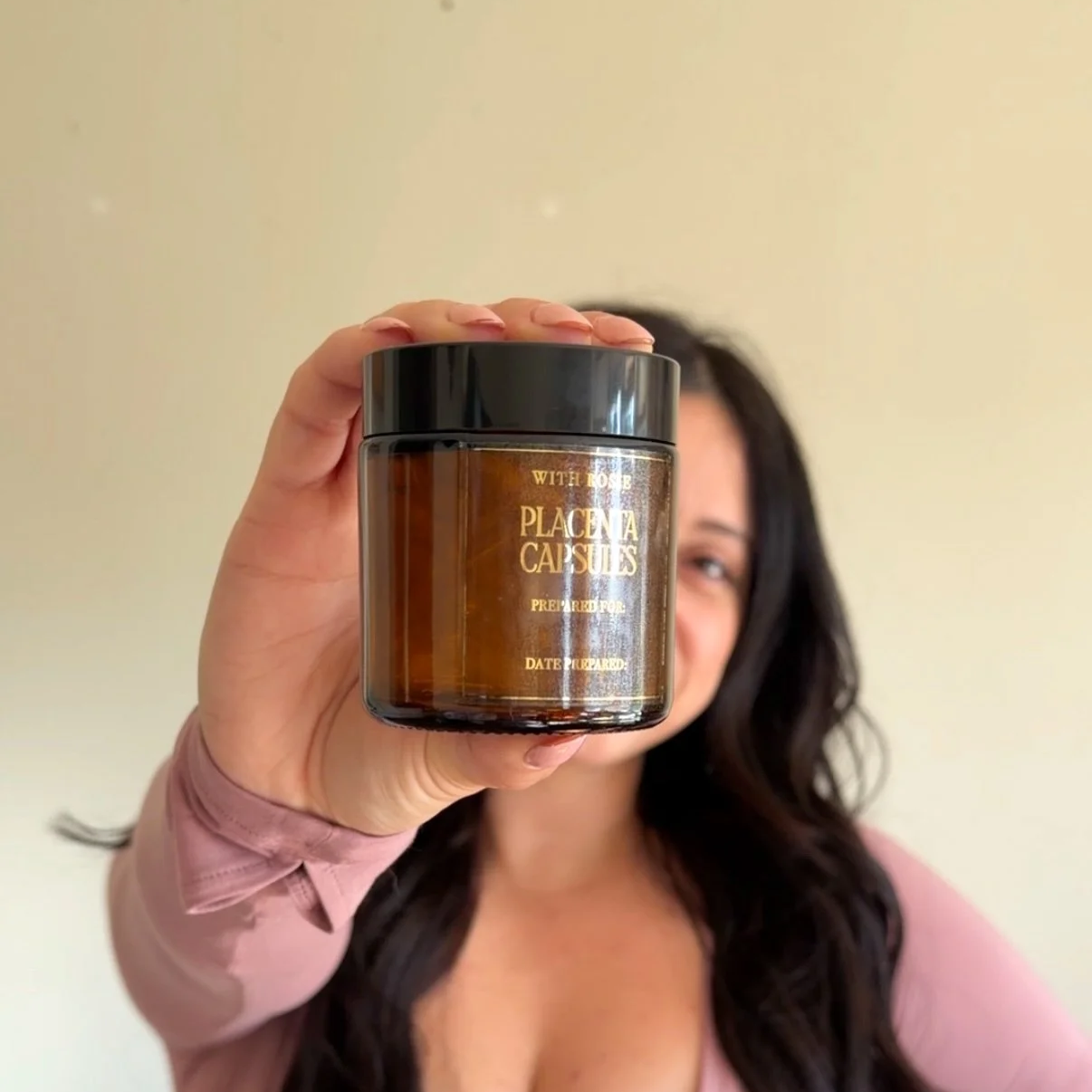 Woman holding a brown jar with gold text on it in front of her face, background is plain off-white wall.