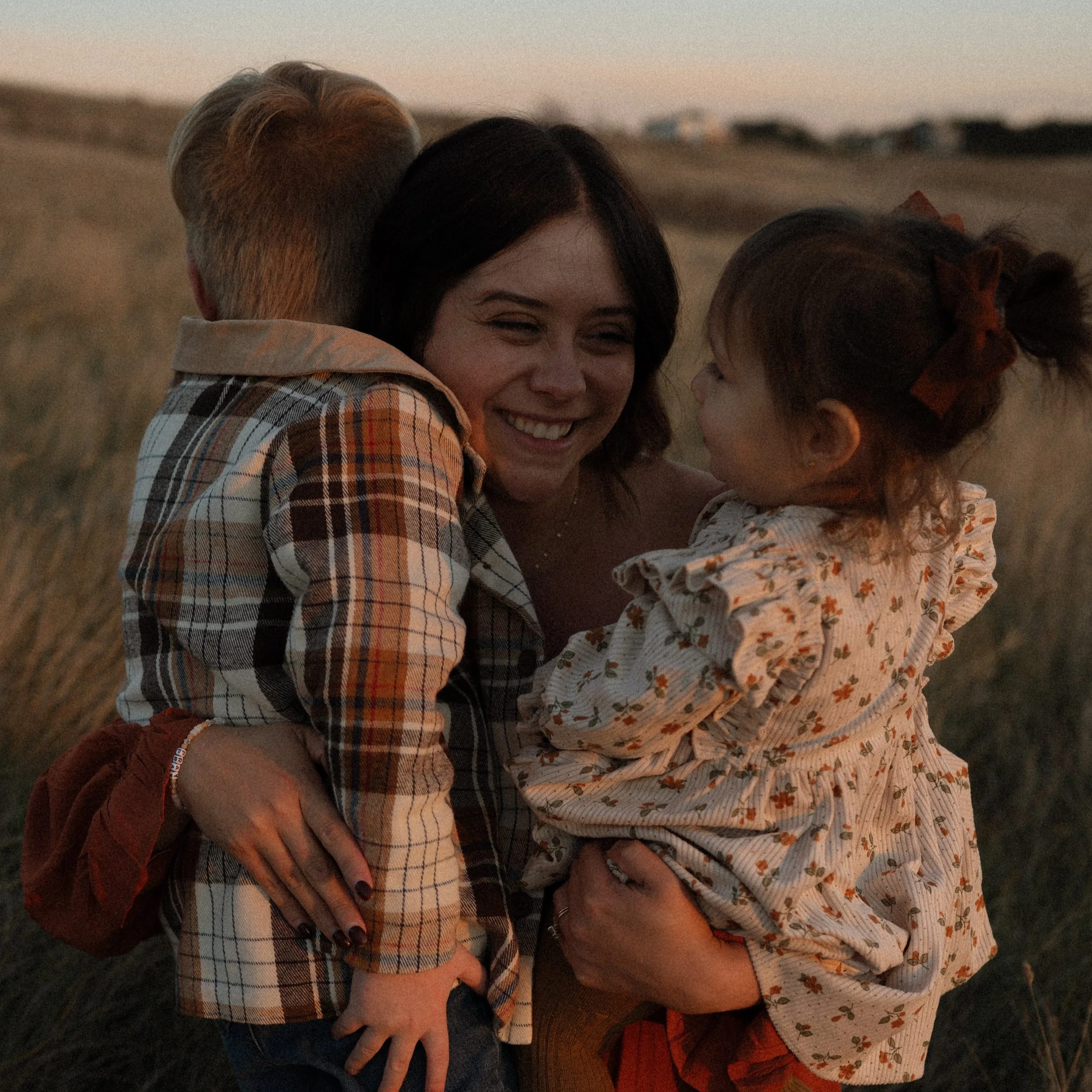 A woman and a young girl share a hug outdoors during sunset, with fields in the background.