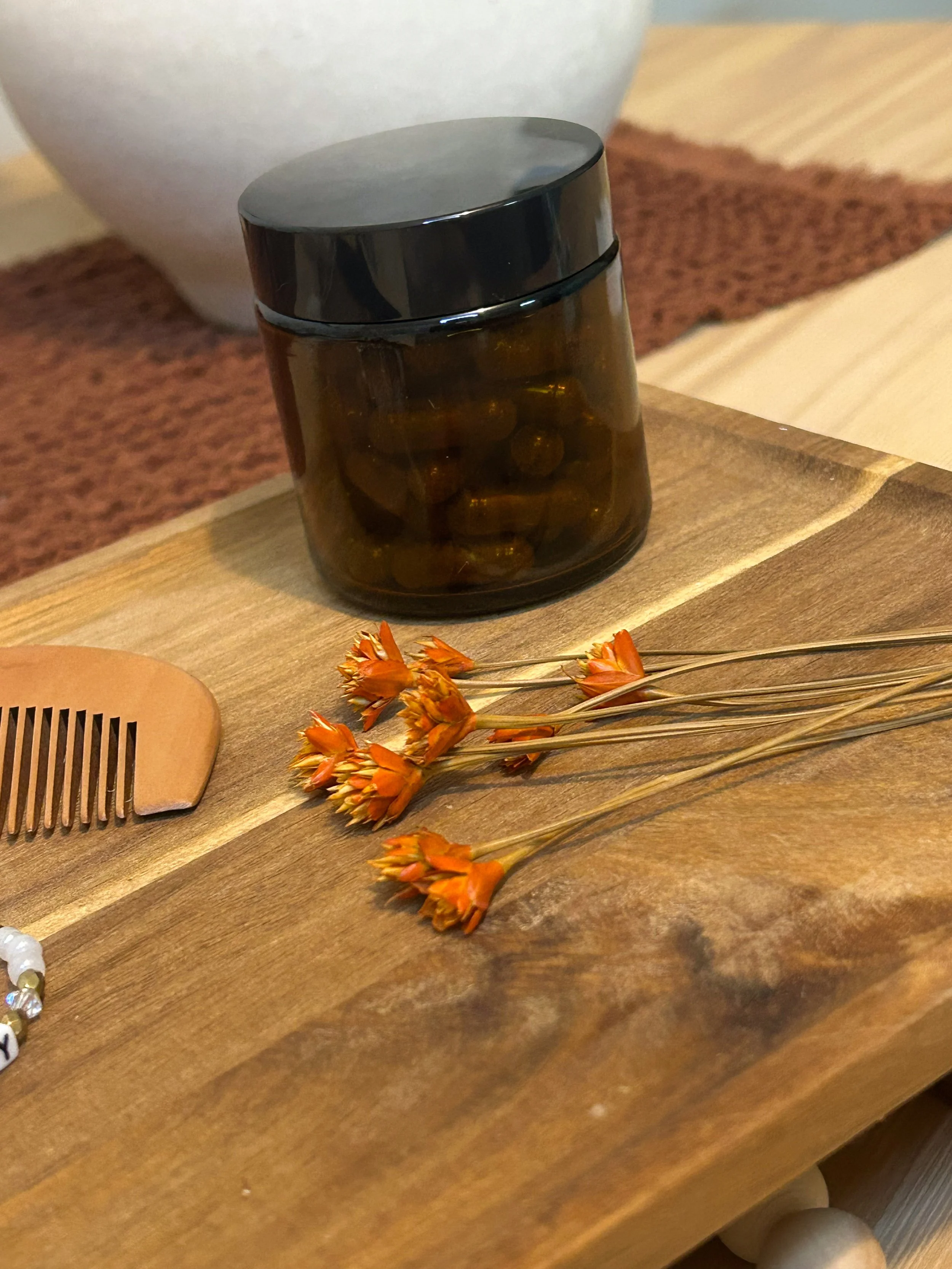 A small dark brown jar with a shiny black lid filled with capsules, placed on a wooden surface next to dried orange flowers.