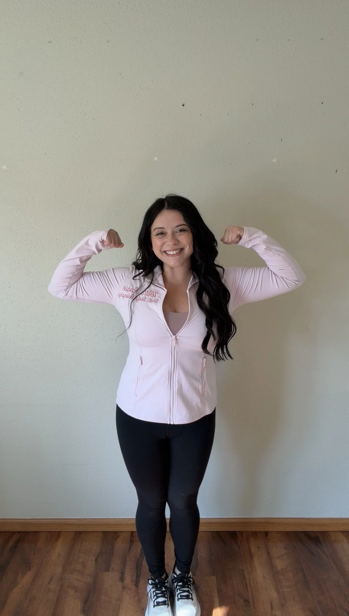 Woman smiling and flexing her biceps while wearing workout clothes and standing on hardwood floor against a plain wall.