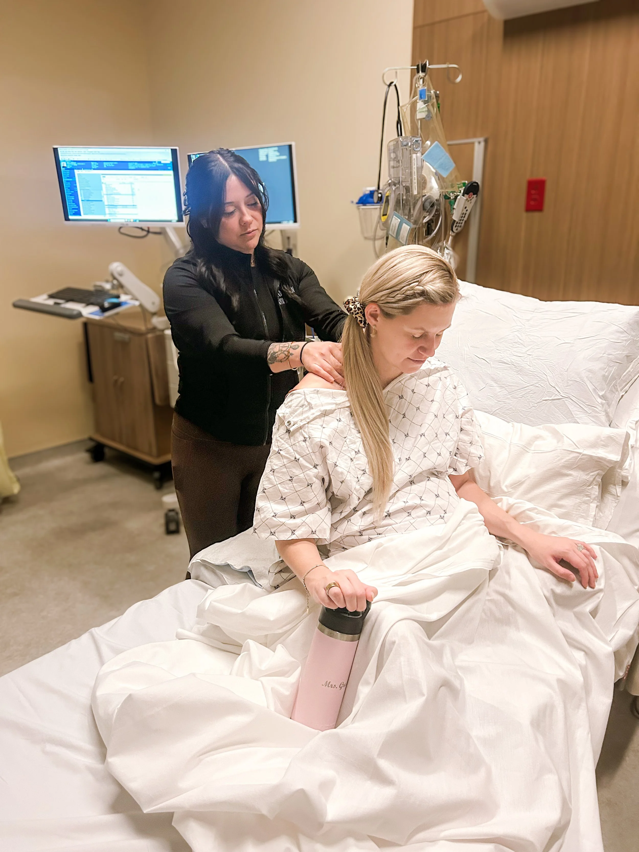 A woman in hospital bed with an IV line receiving a massage from a healthcare worker in a hospital room.