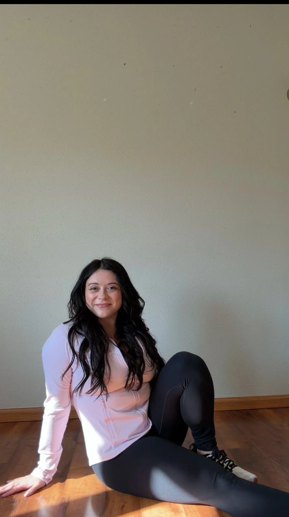 A woman with long black hair, wearing a light pink jacket and black leggings, is sitting on a wooden floor against a plain beige wall, smiling at the camera.