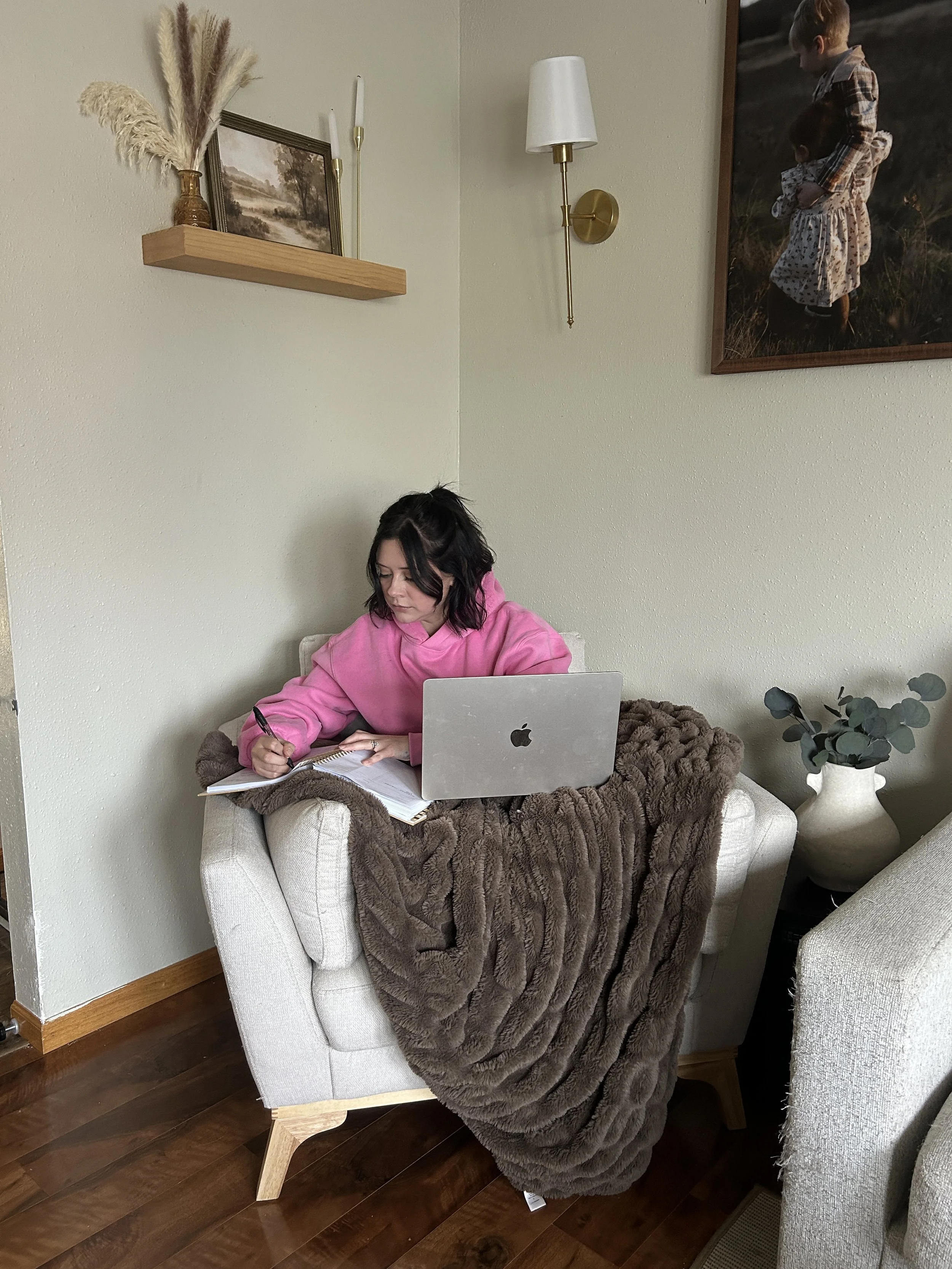 A woman with curly dark hair wearing a pink hoodie sitting on a beige armchair with a laptop and notepad, a brown fur blanket, and a plant with grayish leaves nearby. The background has a wall with wall-mounted lamps, a shelf with a small framed landscape picture, and a large photograph of two children looking at each other, dressed in patterned clothing.