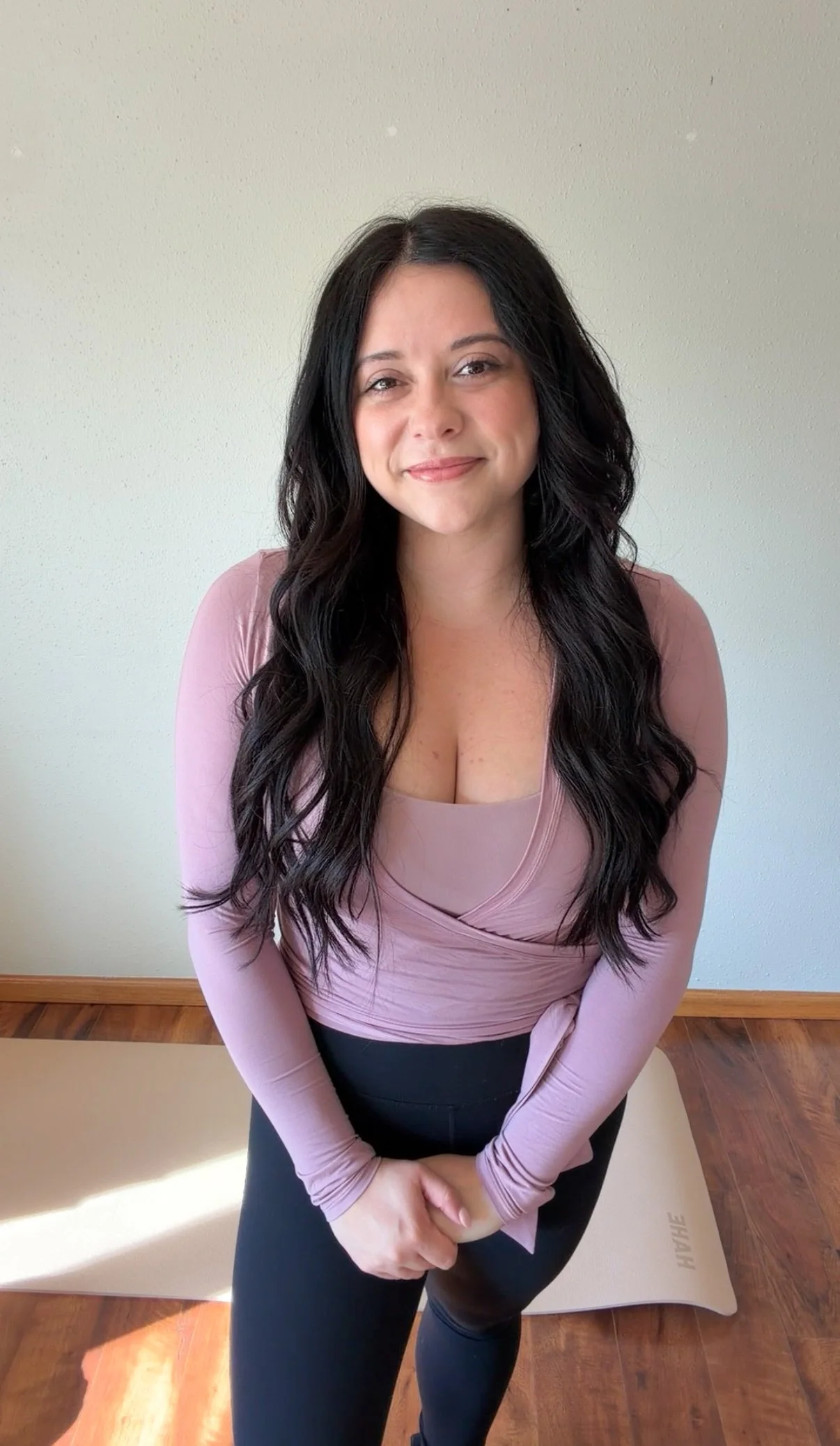 Woman with long dark hair wearing pink long-sleeve top and black pants, standing indoors on wood floor with plain light-colored wall behind her.