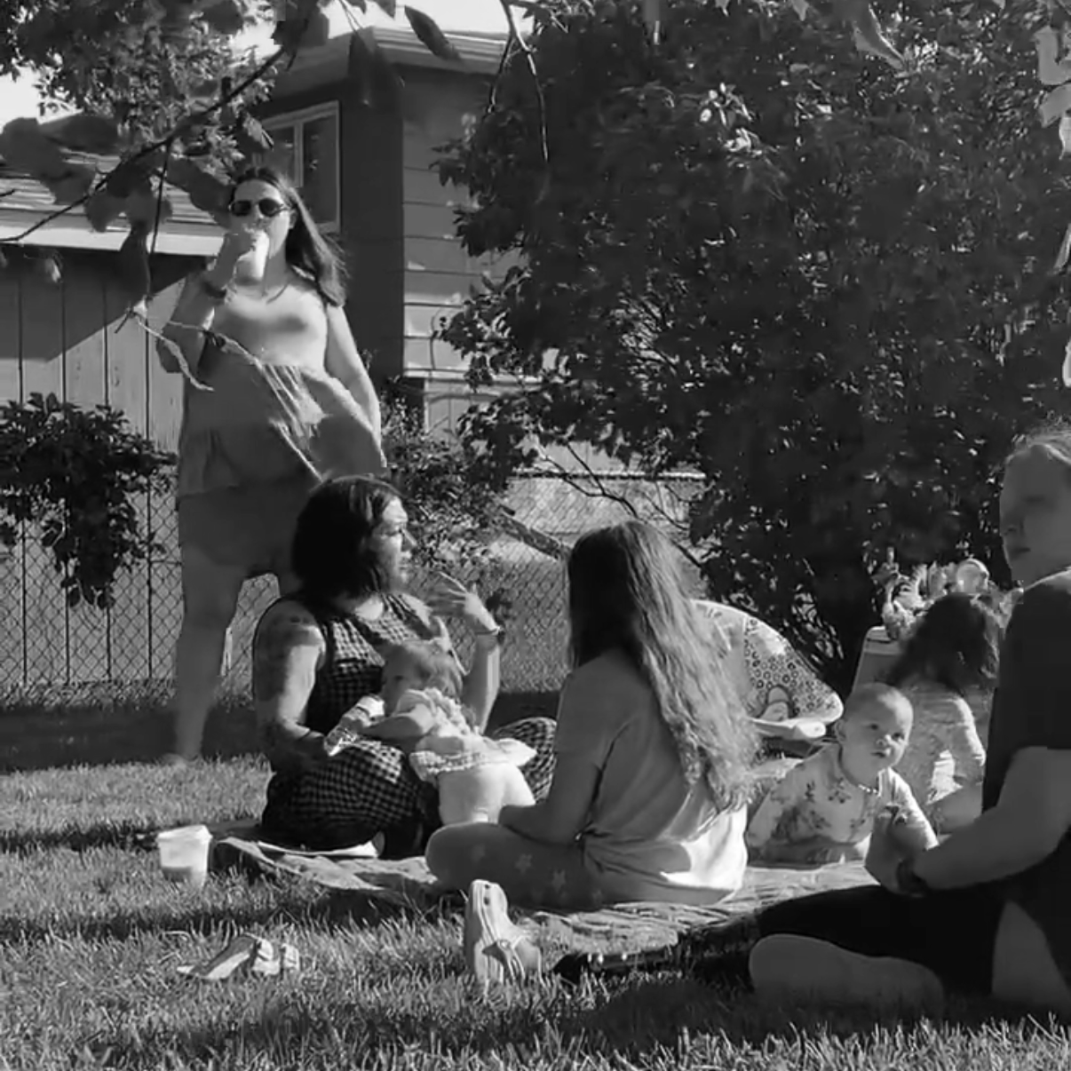 People sitting on a blanket and standing outdoors in a yard, with children and adults, under a tree, with a house and a fence in the background.