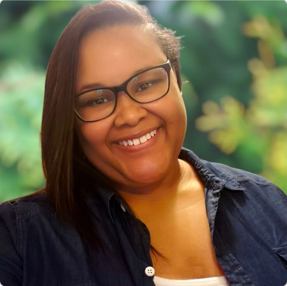 A woman with glasses smiling in front of a blurred green background.