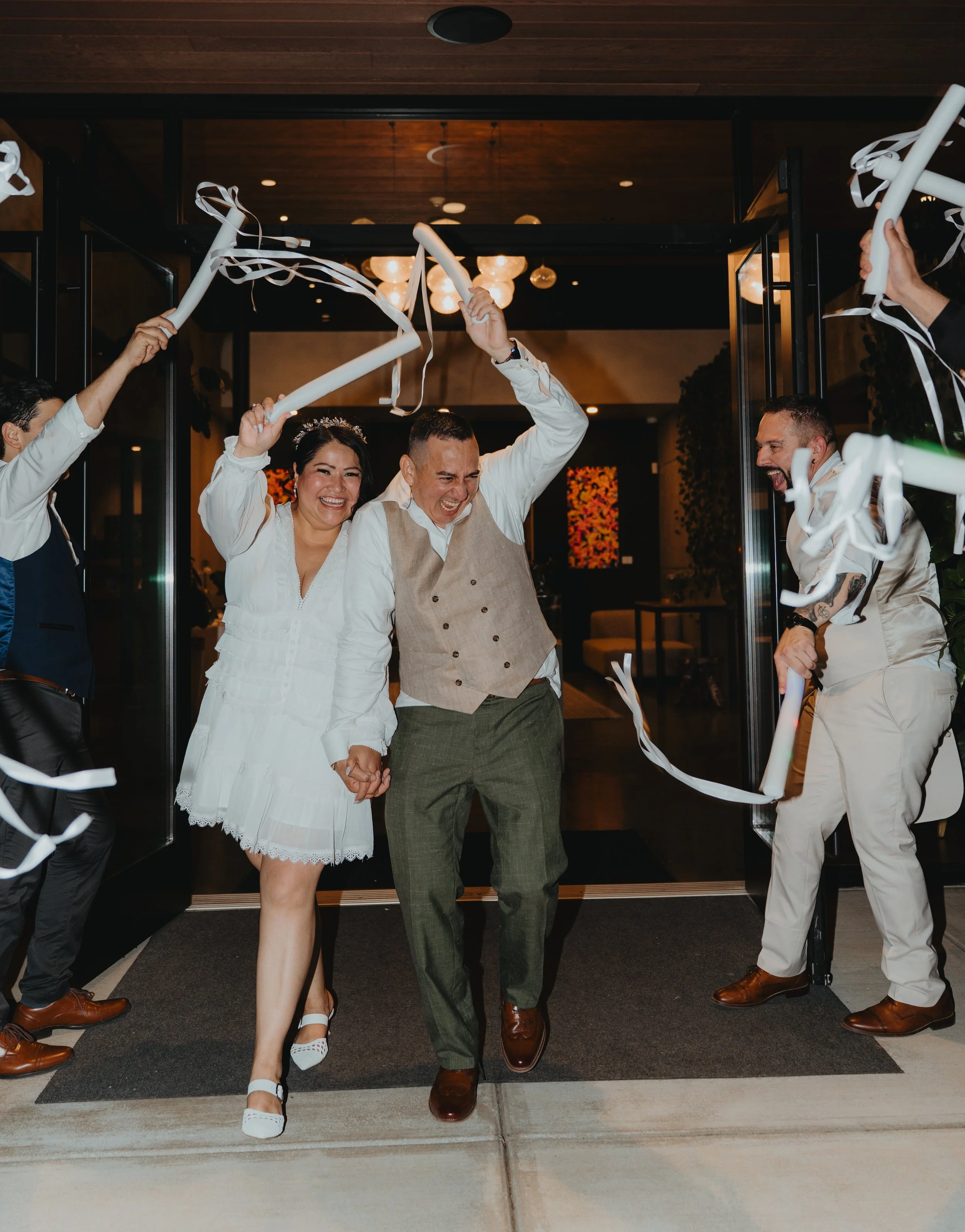 A newlywed couple holding hands and smiling as they walk through a doorway. They are surrounded by friends or family celebrating with white streamers, in an indoor setting with warm lighting.