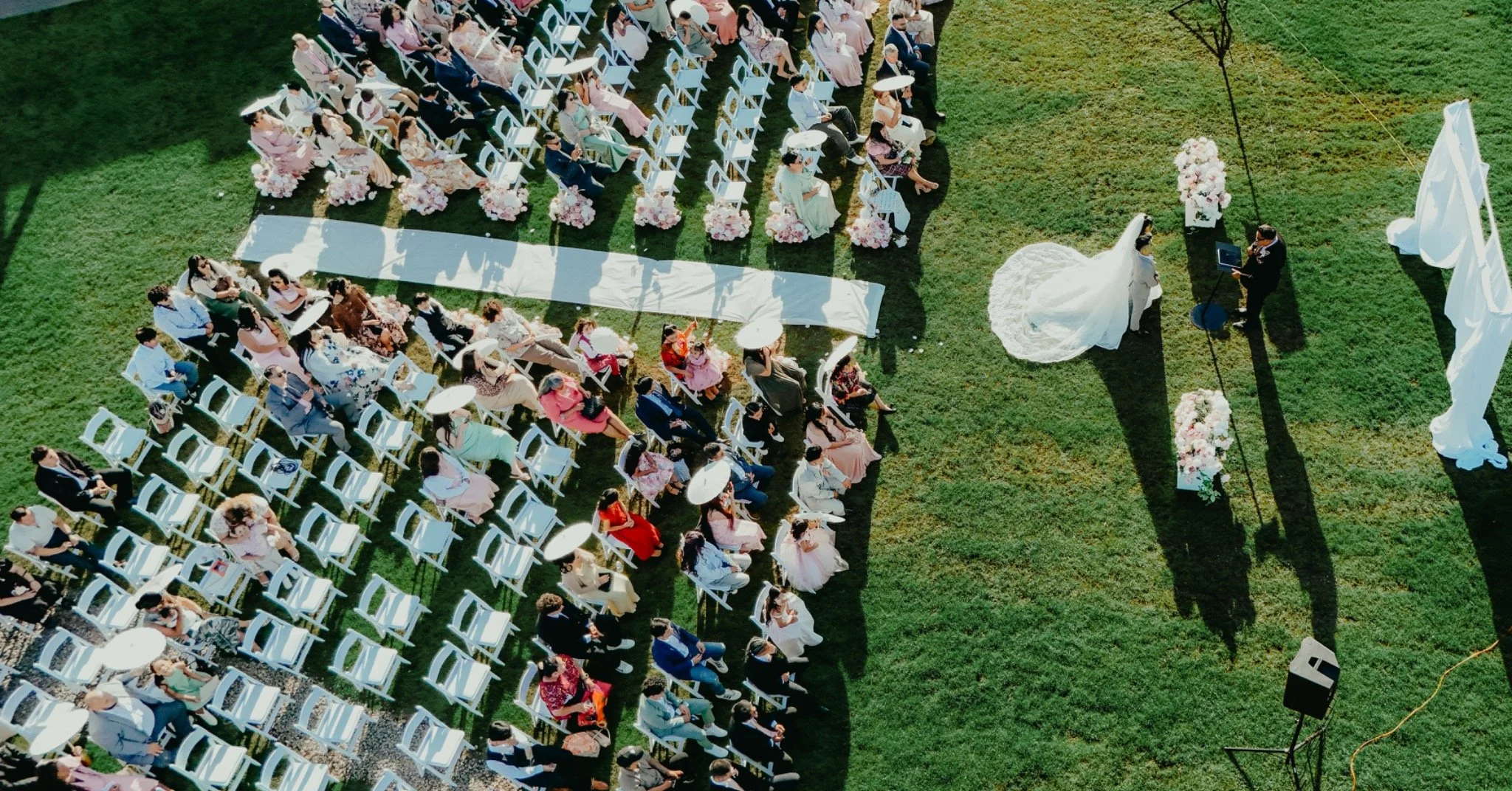 An aerial view of a wedding ceremony outdoors on a grassy field. The bride and groom are standing near an altar decorated with flowers, with guests seated in white chairs arranged in rows. Some guests are holding parasols, and the bride is wearing a white gown while the groom is in a dark suit.