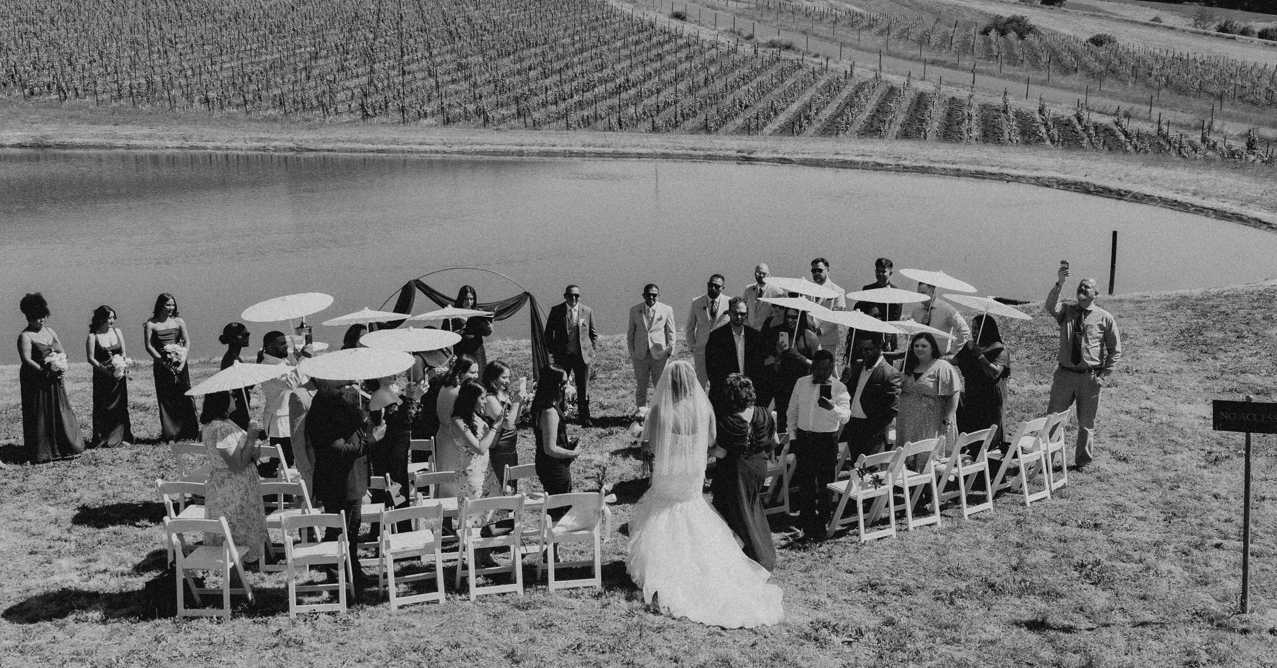 A wedding ceremony taking place outdoors near a body of water with vineyard hills in the background. The bride and groom are standing in front of seated guests, with guests holding umbrellas for shade. The scene is in black and white.