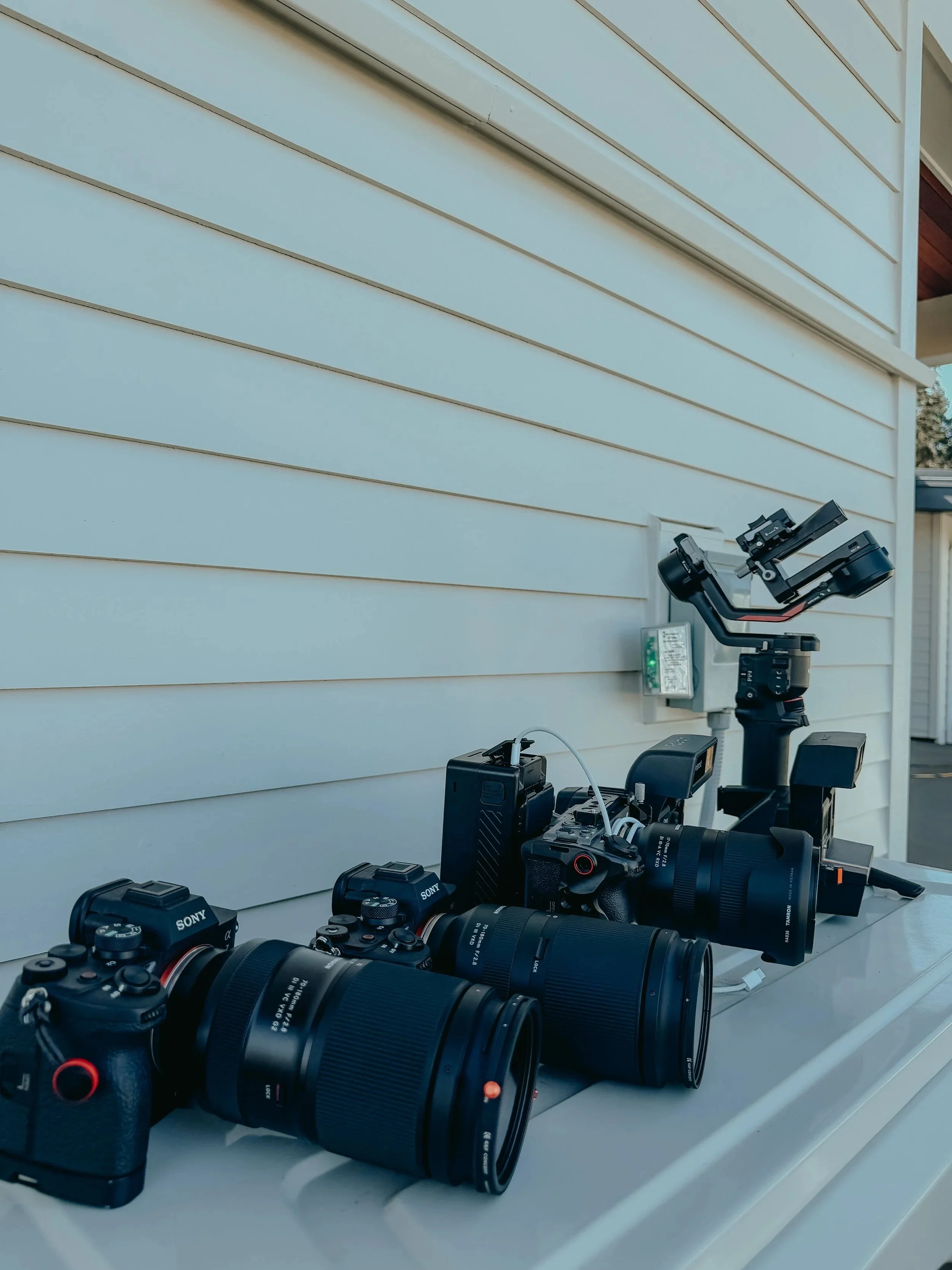 A collection of camera equipment including professional cameras and lenses arranged on a white surface outdoors, with a light-colored building side wall in the background.