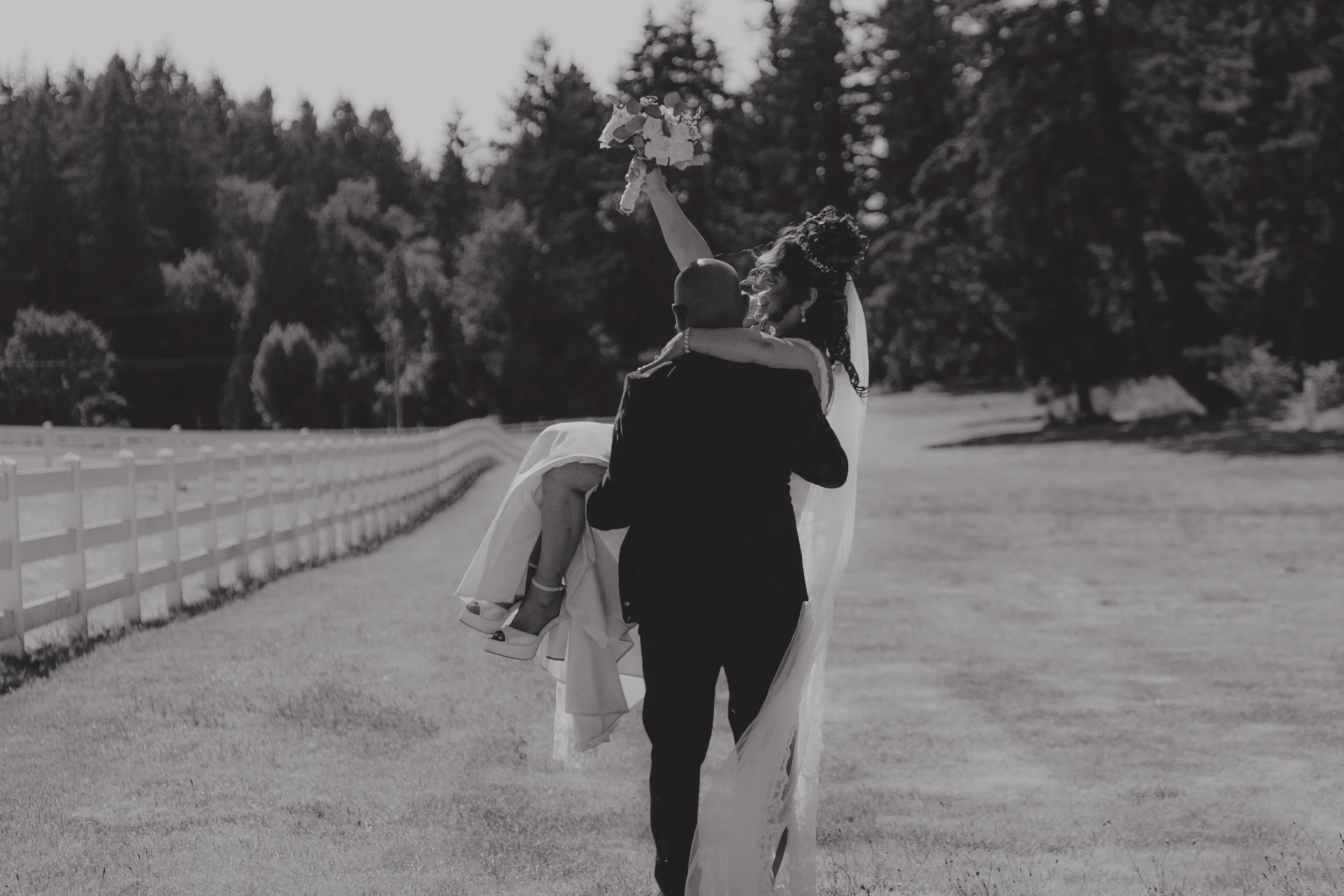 A bride and groom dancing outdoors, with the groom lifting the bride in a white wedding dress. The bride is holding a bouquet and smiling. There is a white fence and trees in the background.
