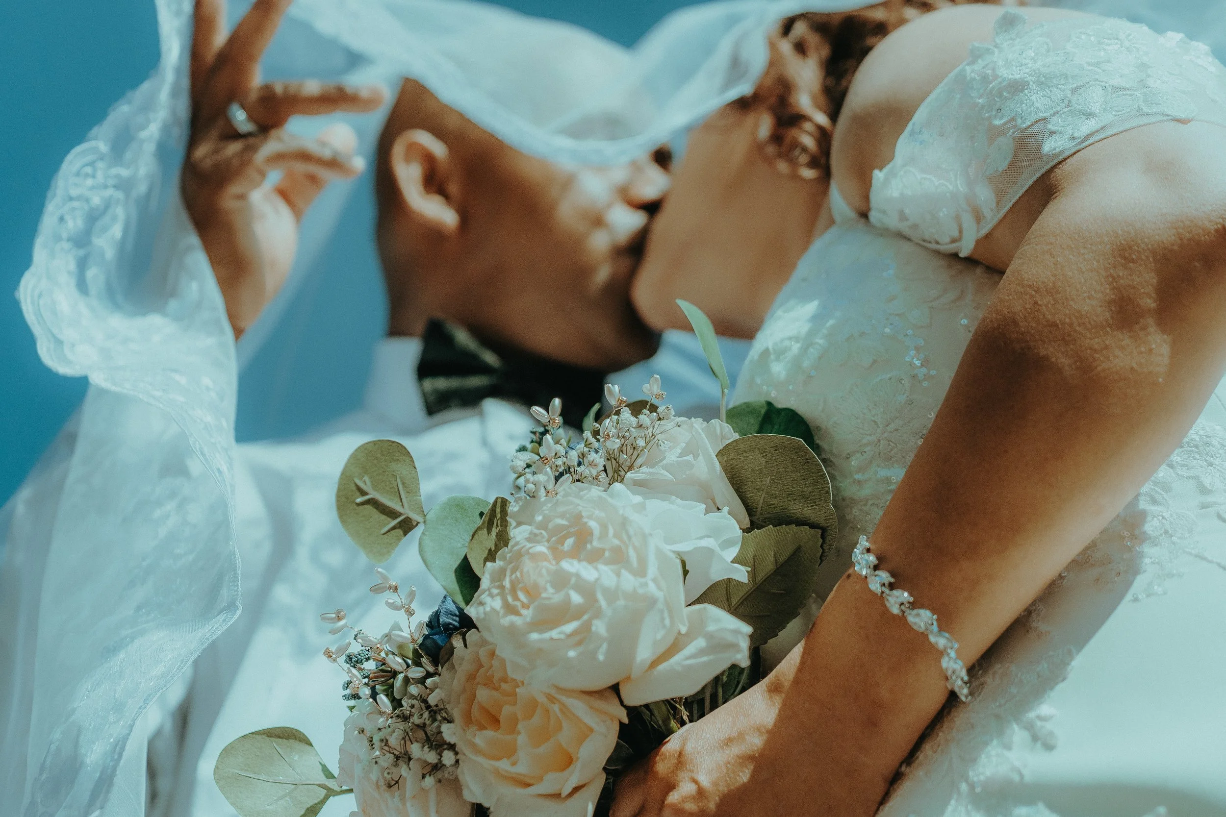 Close-up of a bride in a wedding dress with lace details, holding a bouquet of cream and white flowers with green leaves, while slightly leaning forward.