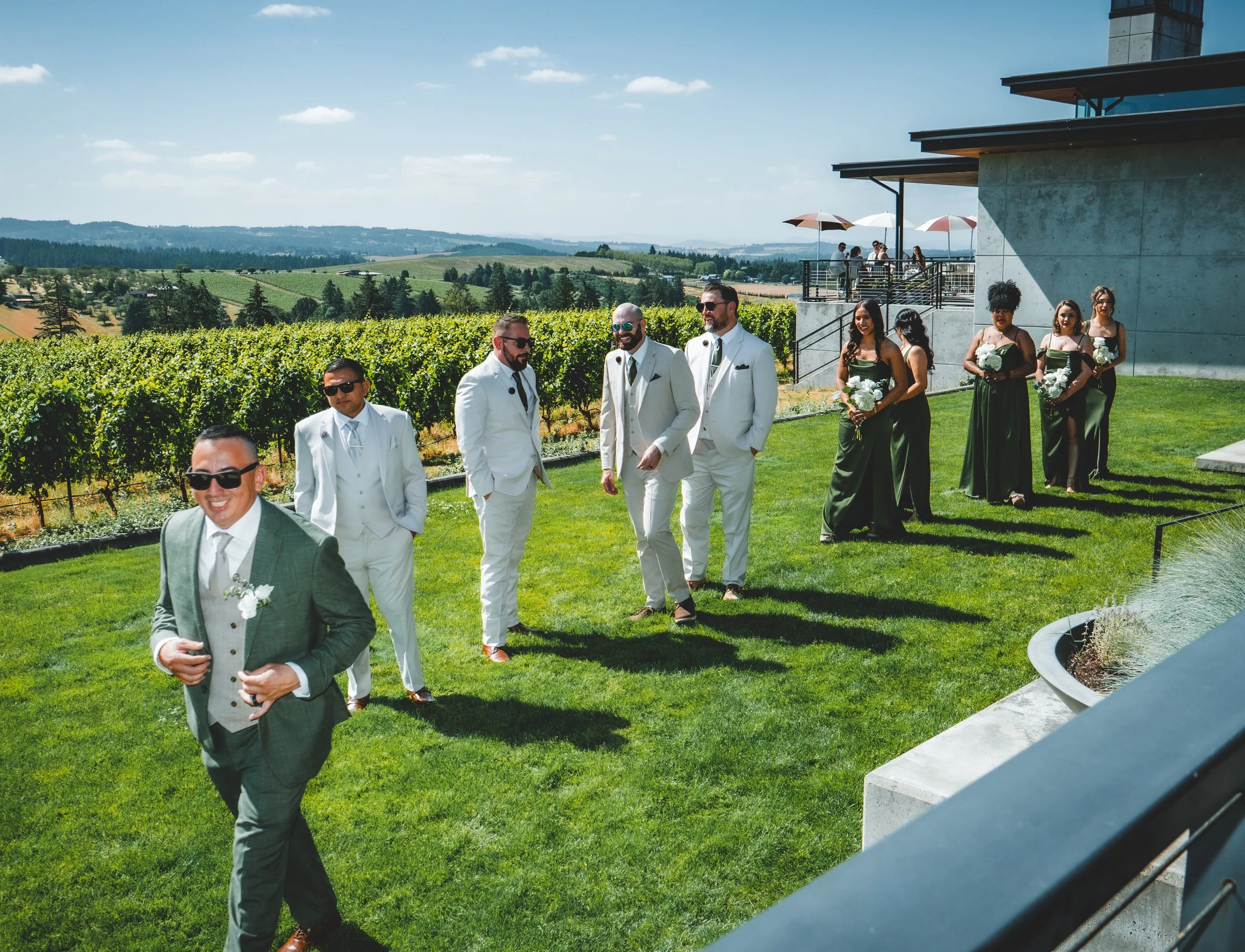 A wedding party walking outdoors on a sunny day in a rural vineyard setting. The group includes men in white suits and women in green dresses, some holding bouquets. A modern building with an outdoor balcony is visible in the background, along with scenic hills and vinyard fields.