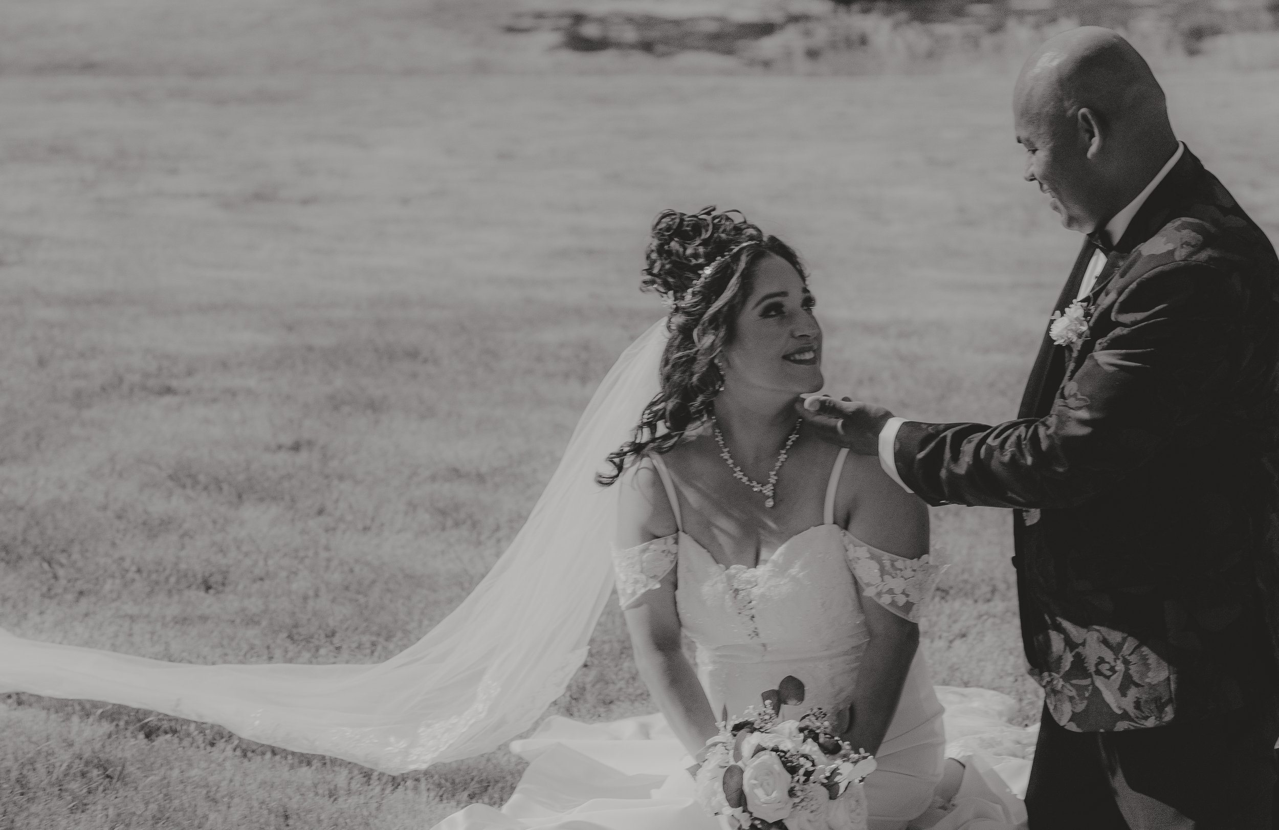 A black and white photo of a bride and groom at a wedding, standing outdoors on a grassy field. The bride is looking up at the groom with a smile, wearing a lace wedding dress and a veil, holding a bouquet of flowers. The groom, dressed in a suit, is looking down at her with a smile, holding her hand gently.